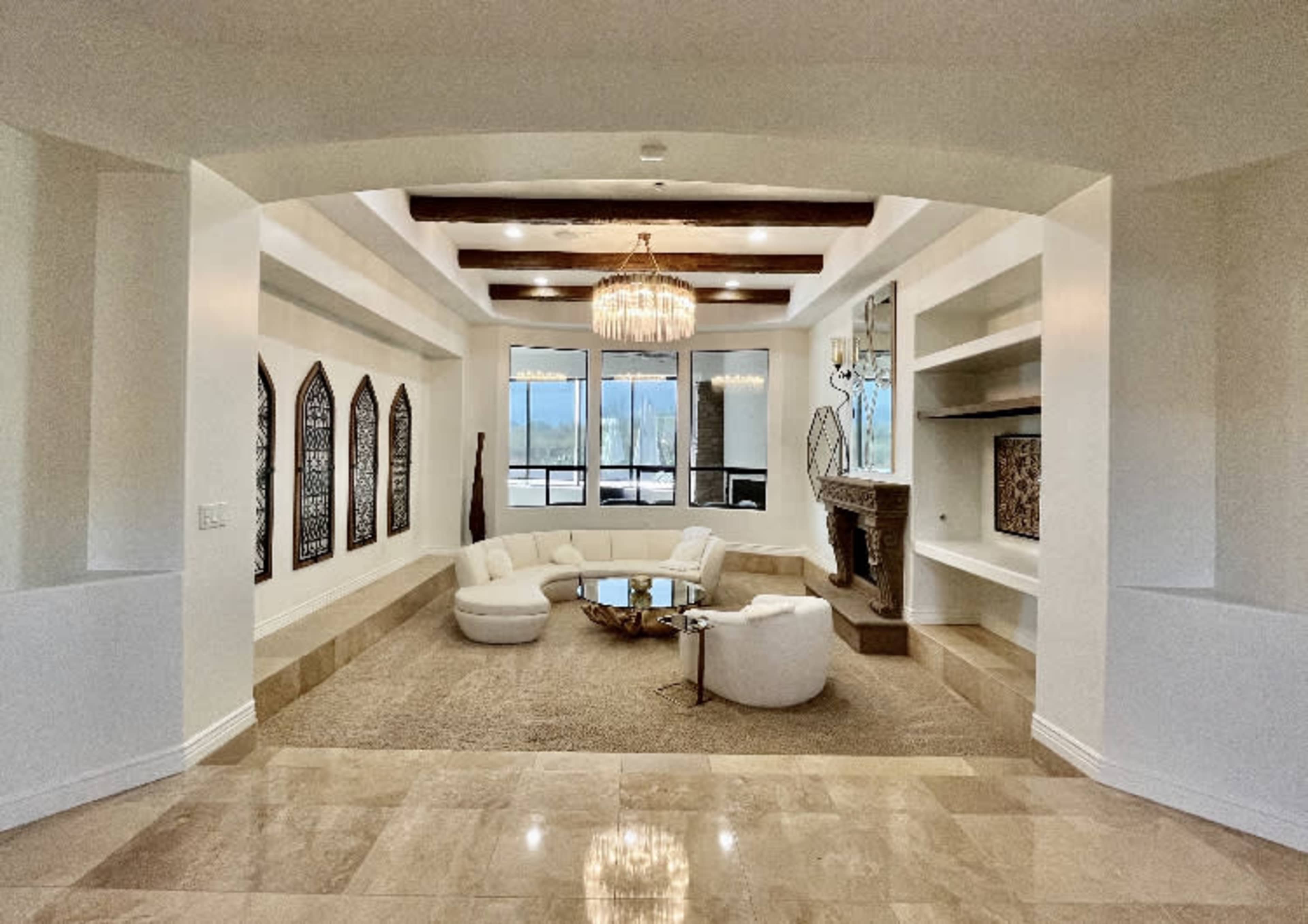 The image shows a spacious living room with a circular seating arrangement, a chandelier, large windows, and a stone fireplace.