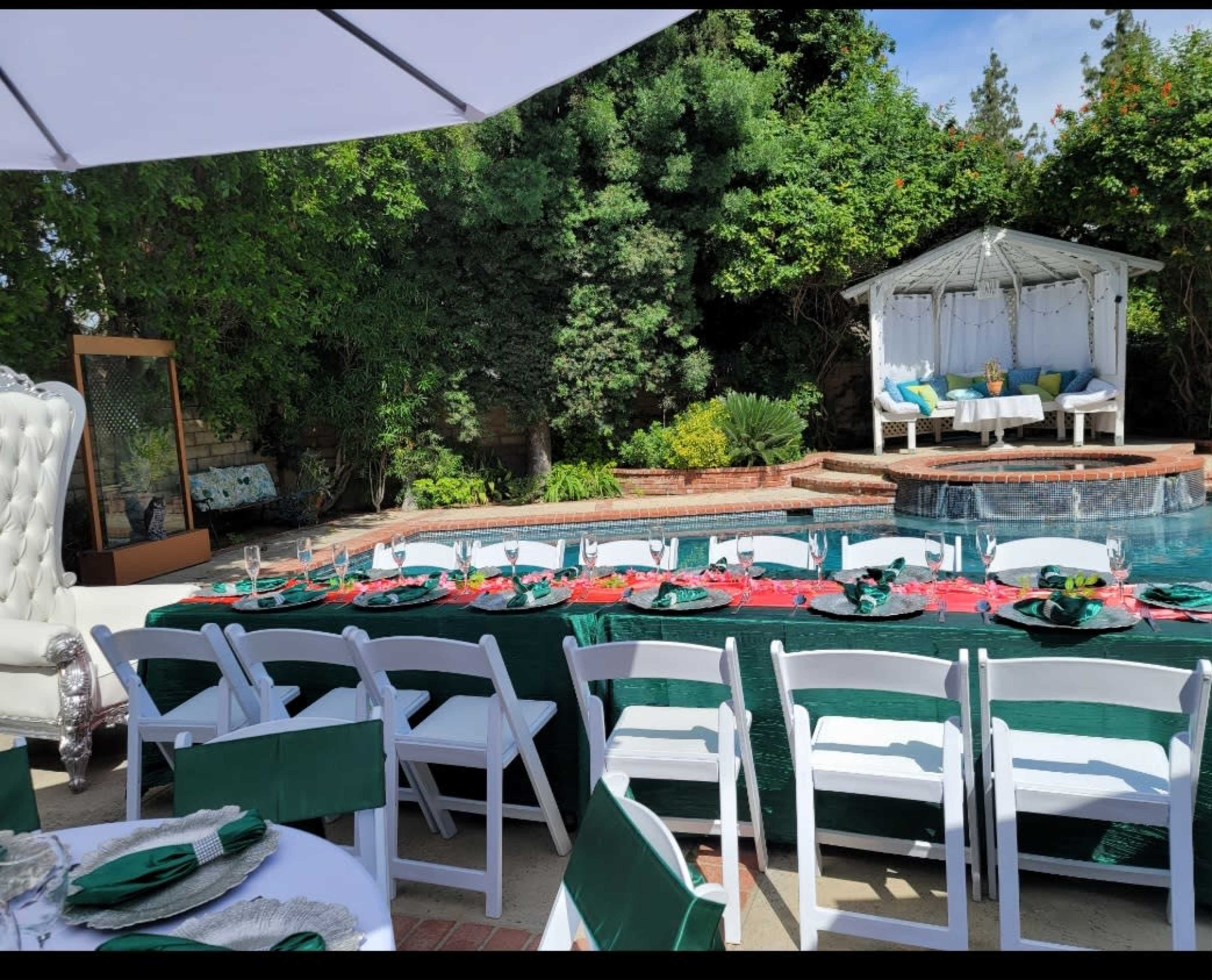 A dining setup with white chairs and a green tablecloth is arranged beside a swimming pool, with a gazebo in the background and greenery surrounding the area.