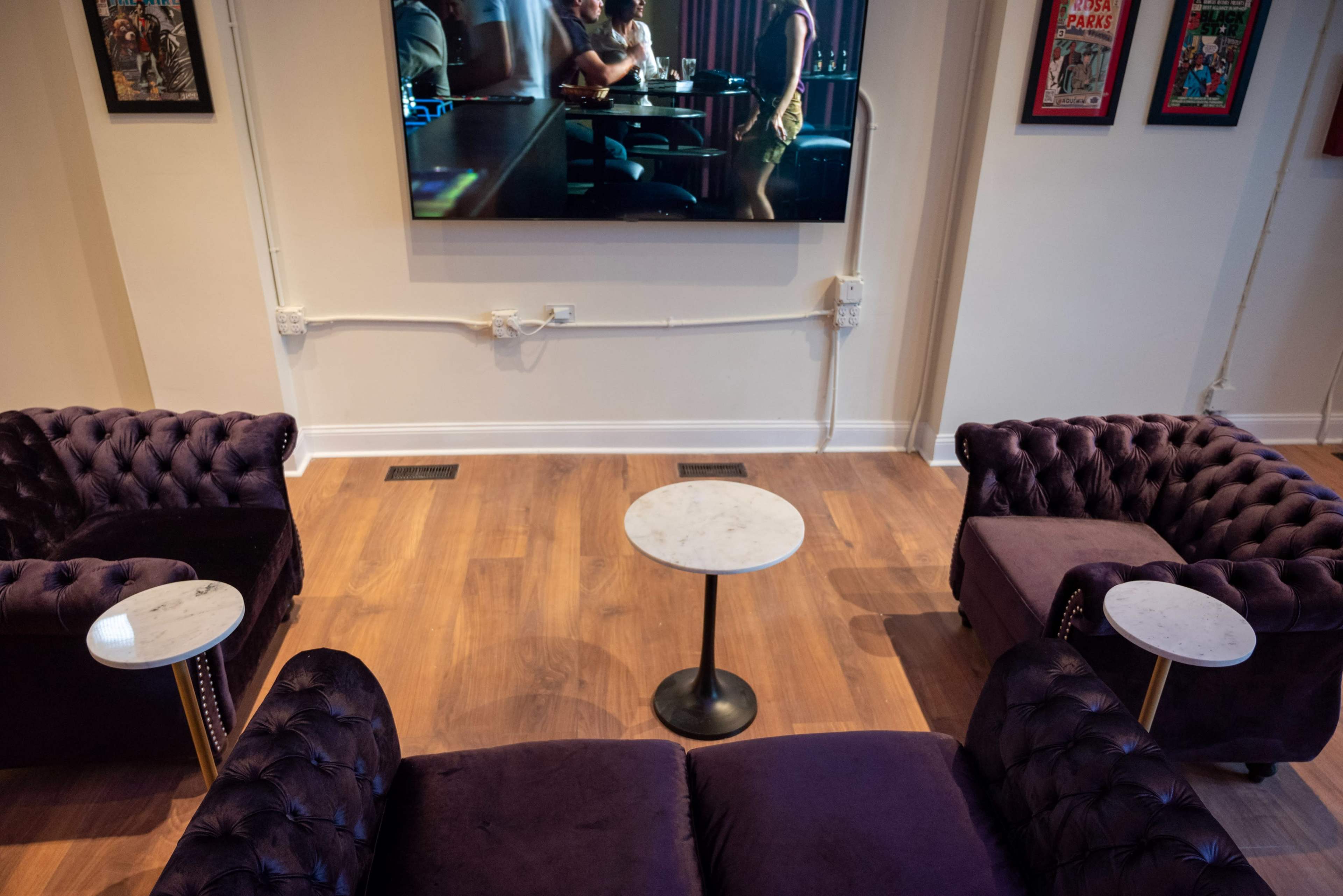 A stylish seating area with two purple velvet sofas and small round tables facing a large television mounted on the wall.