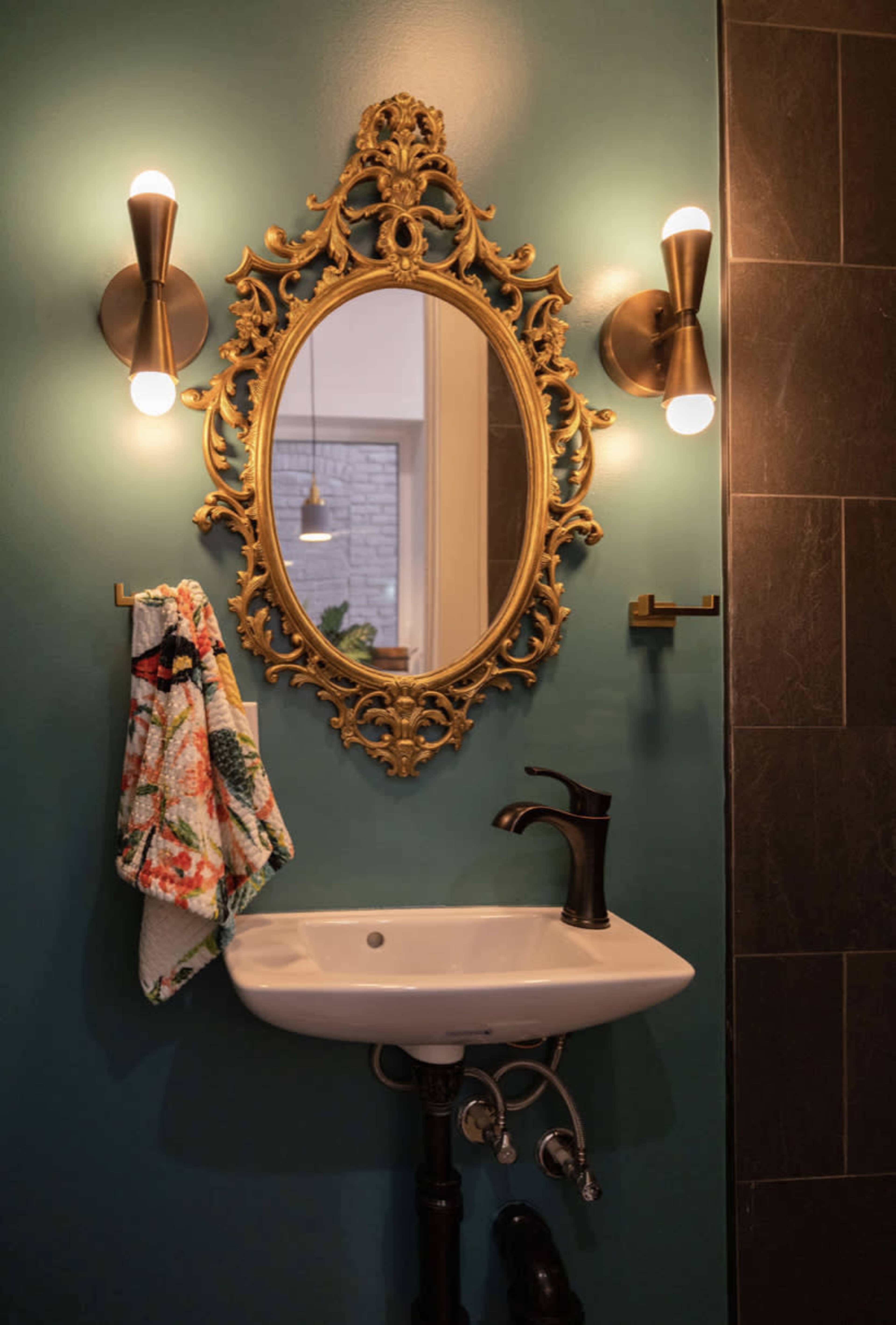 A decorative gold-framed mirror above a white pedestal sink, with two wall-mounted lights flanking the mirror and a colorful towel hanging nearby.