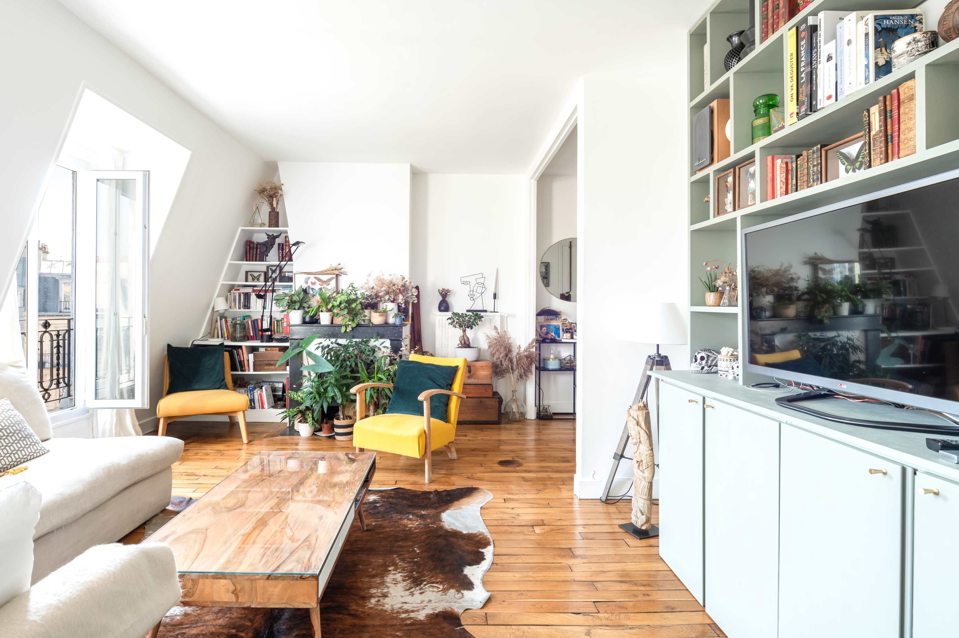 A bright living room features a wooden coffee table, yellow chairs, a bookshelf filled with plants and books, and large windows letting in natural light.