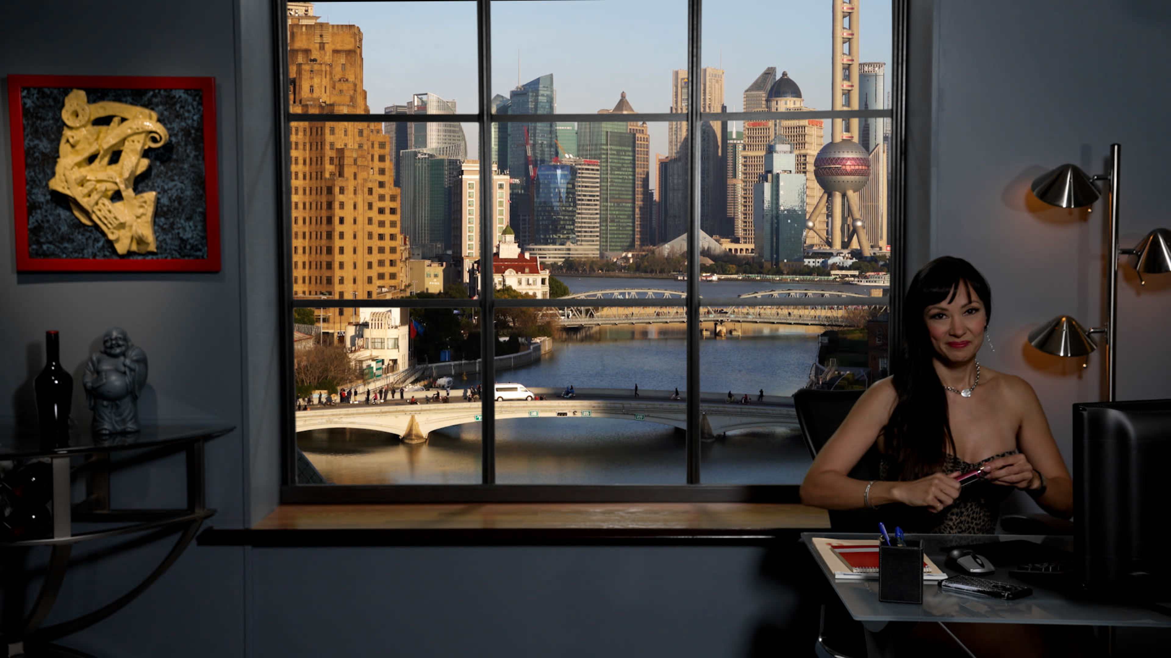 A woman sits at a desk in an office, with a view of a modern city skyline through a large window.