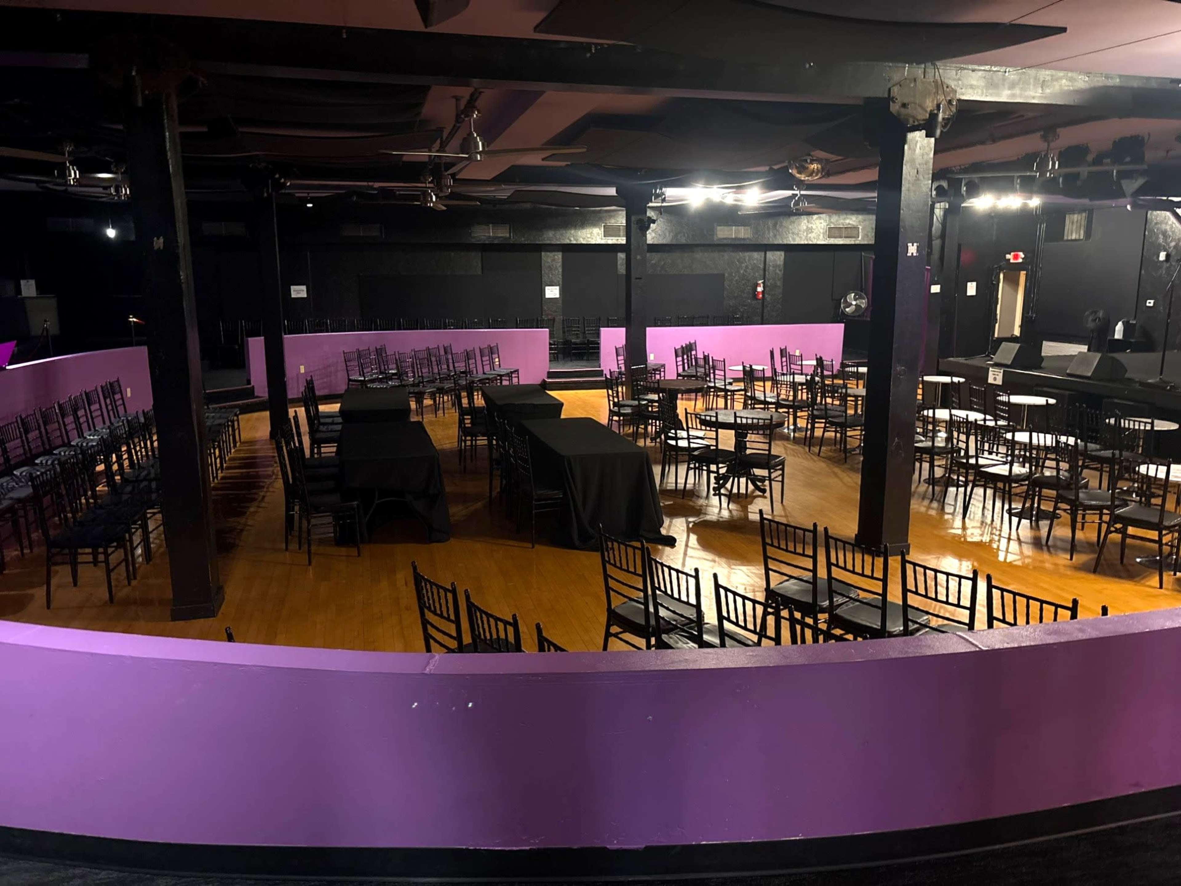 The image shows a performance venue with rows of empty chairs and tables arranged around a central floor area, all under dim lighting.