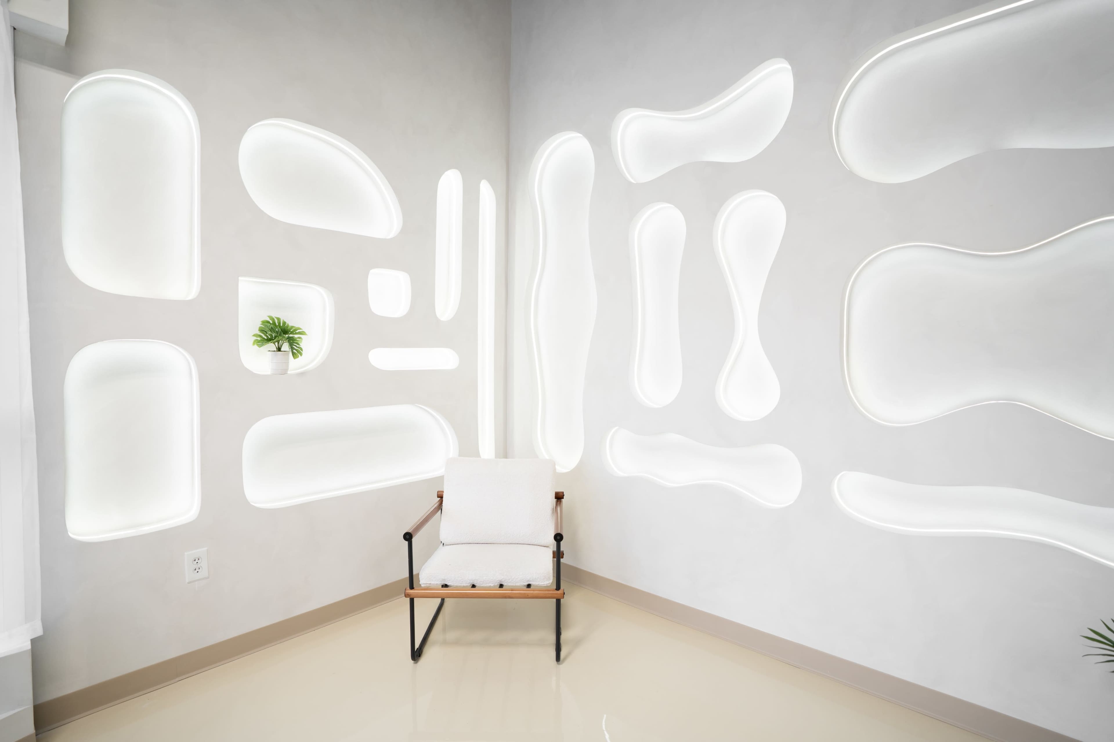 The image shows a minimalist interior with a single chair against a wall featuring illuminated, organic-shaped niches.