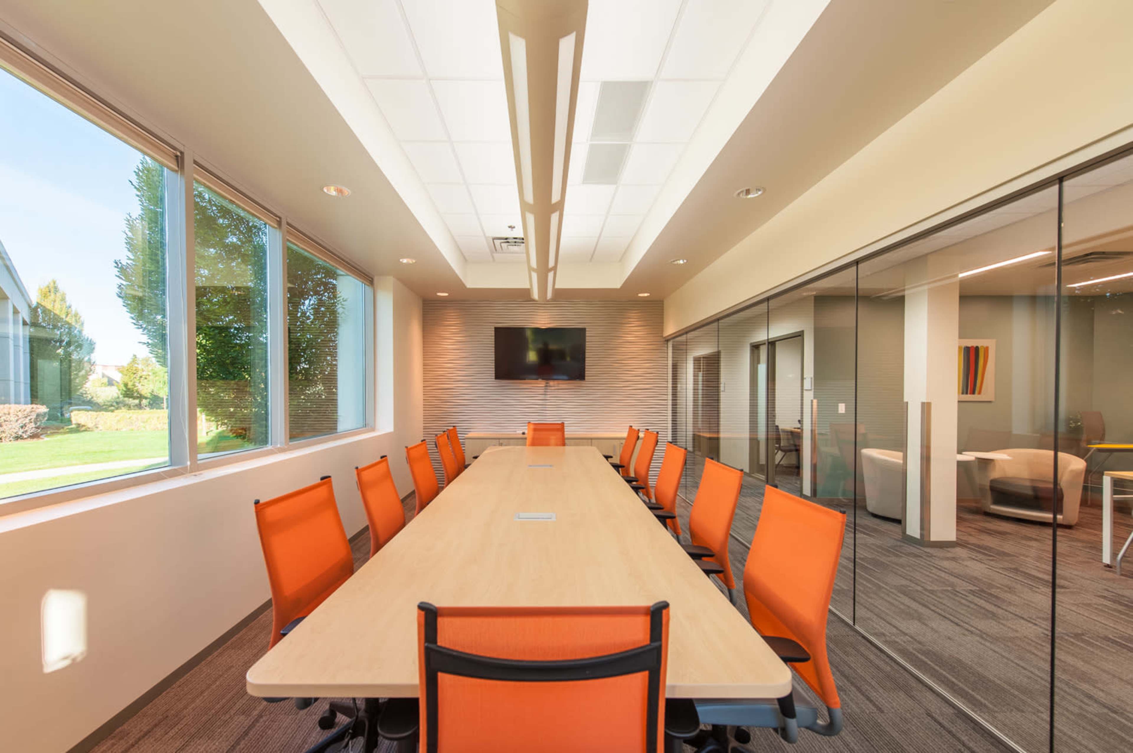 A modern conference room features a long wooden table surrounded by orange chairs, with large windows allowing natural light to fill the space.