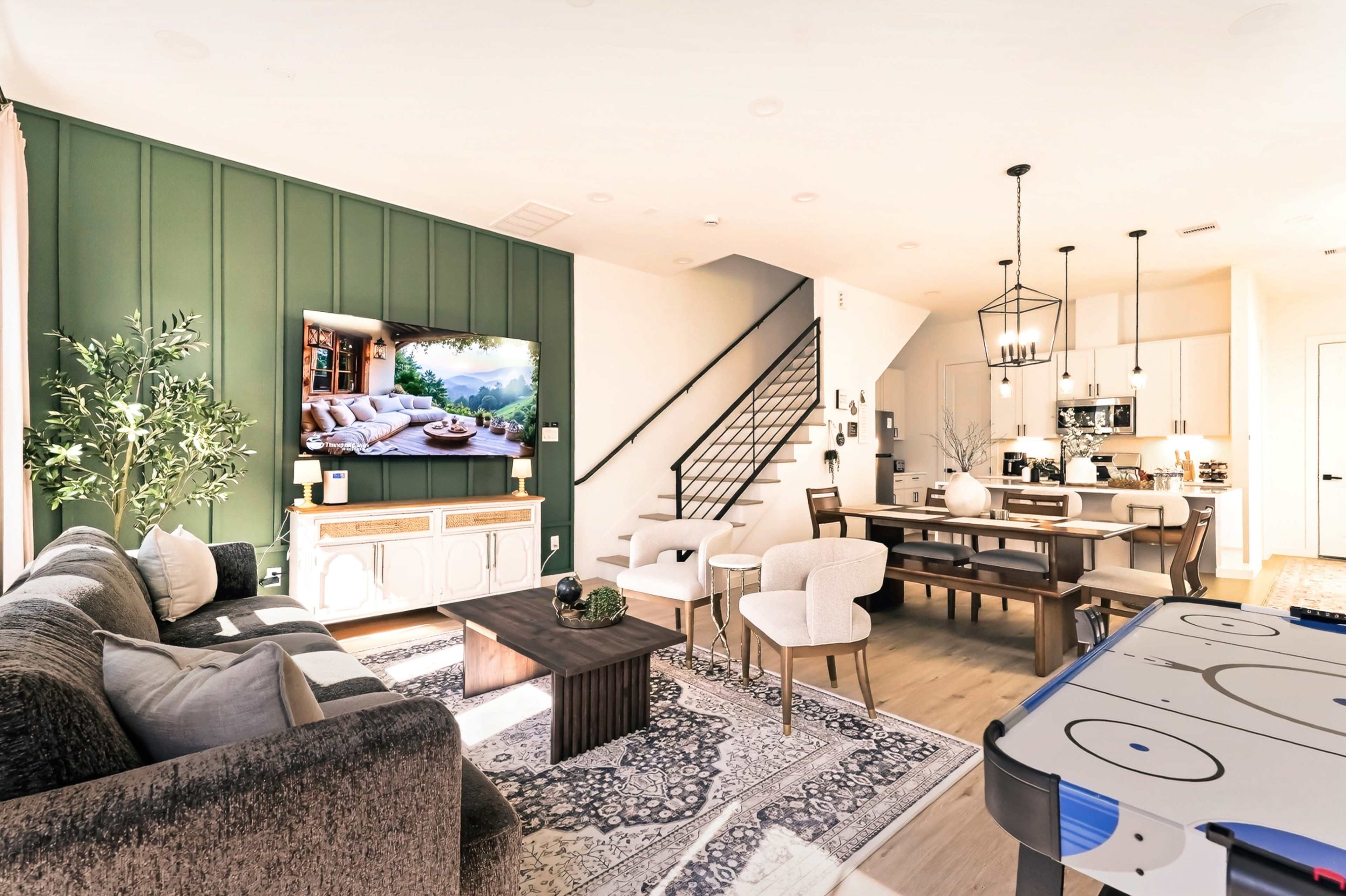 The image depicts a modern living space featuring a seating area with a large TV mounted on a green accent wall, a dining area with a table and chairs, and a staircase leading to an upper level.