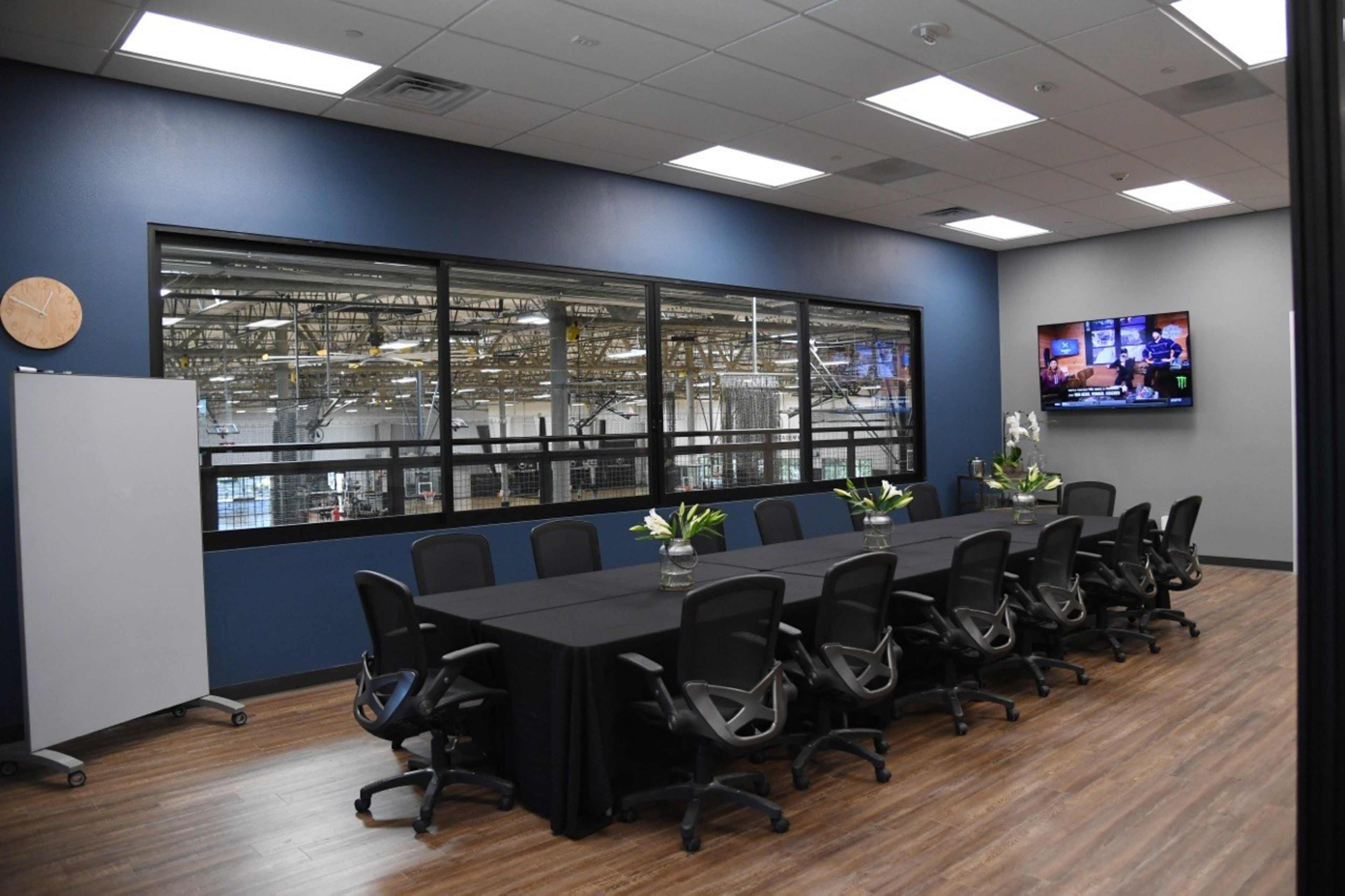 A large conference room features a long table surrounded by office chairs, with a view of a warehouse through large windows and a television mounted on the wall.