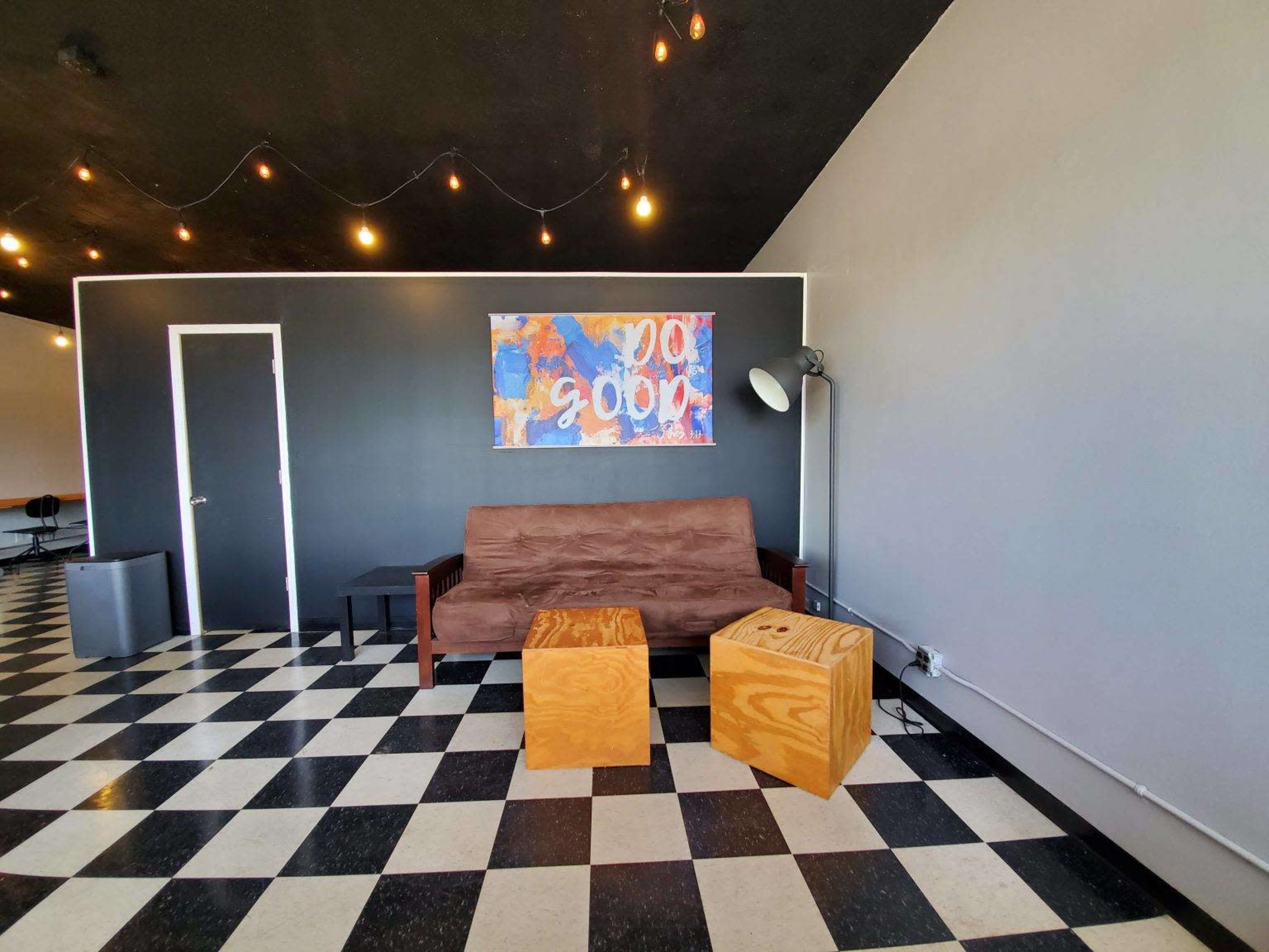 A room with a brown couch, two wooden cube tables, a lamp, and a door, all set against a checkered black and white floor and a gray wall with a colorful painting.