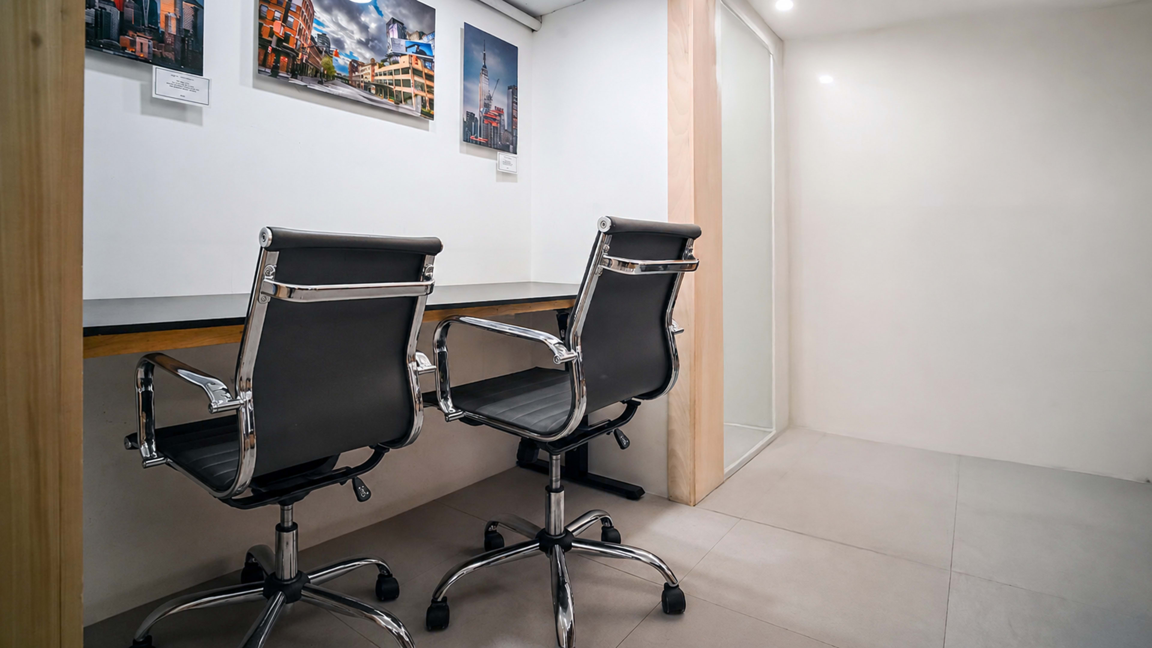 The image shows a modern office space featuring two black swivel chairs facing a wooden desk, with framed artwork on the wall and a glass partition in the background.
