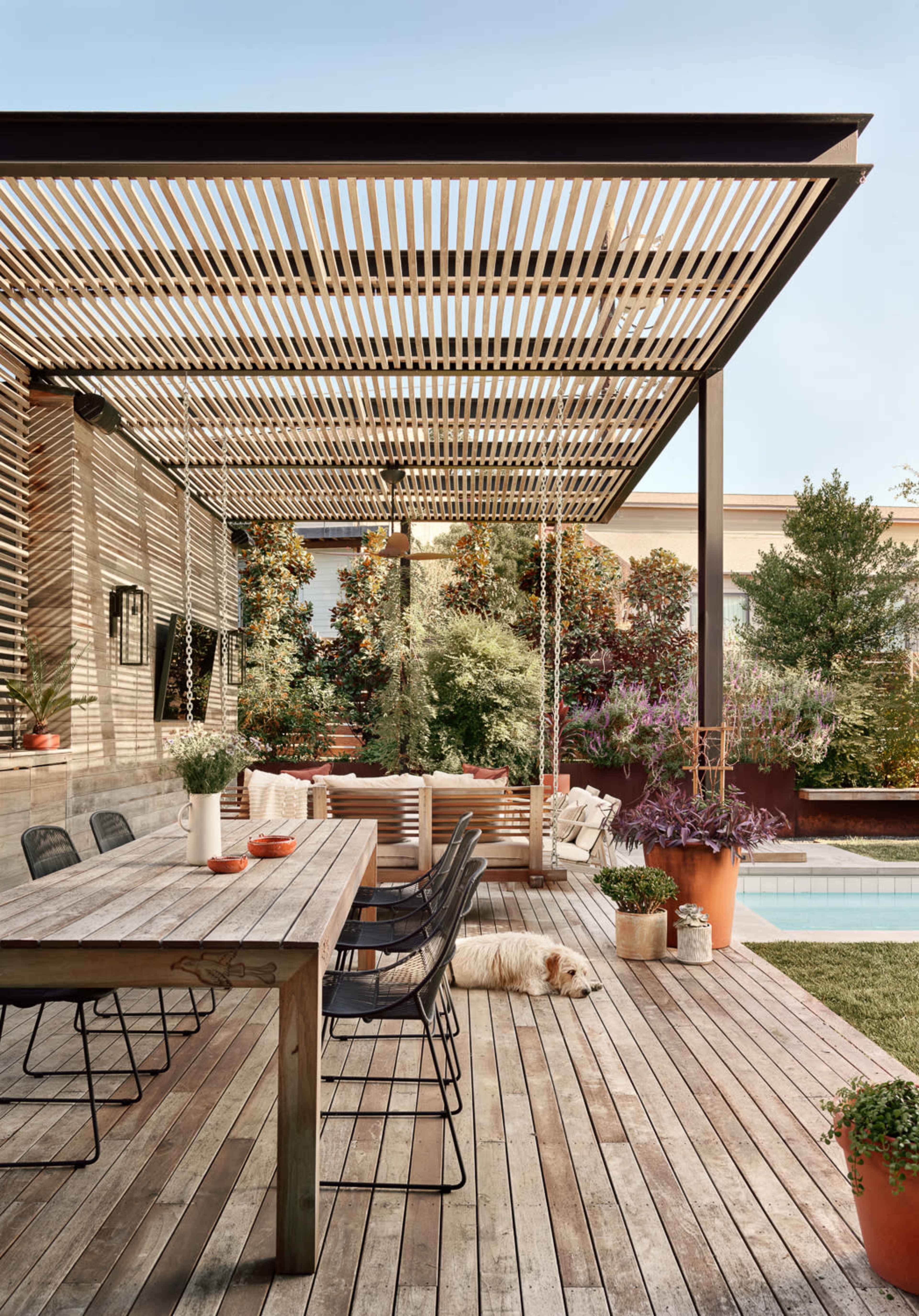 A wooden deck features a large dining table and chairs under a slatted pergola, with potted plants surrounding a swimming pool in the background.
