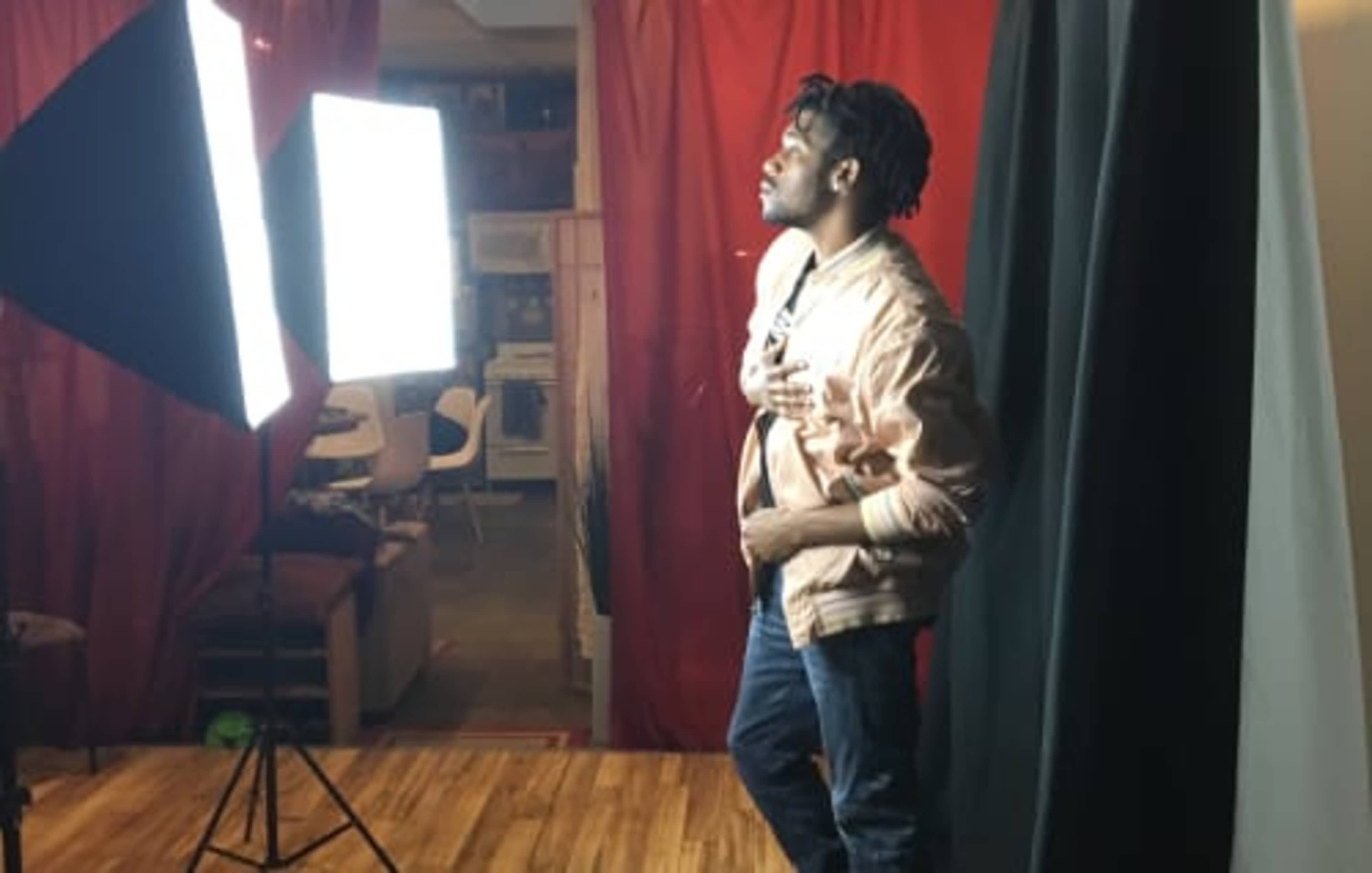 A man stands near two bright studio lights, posing in front of a red backdrop.
