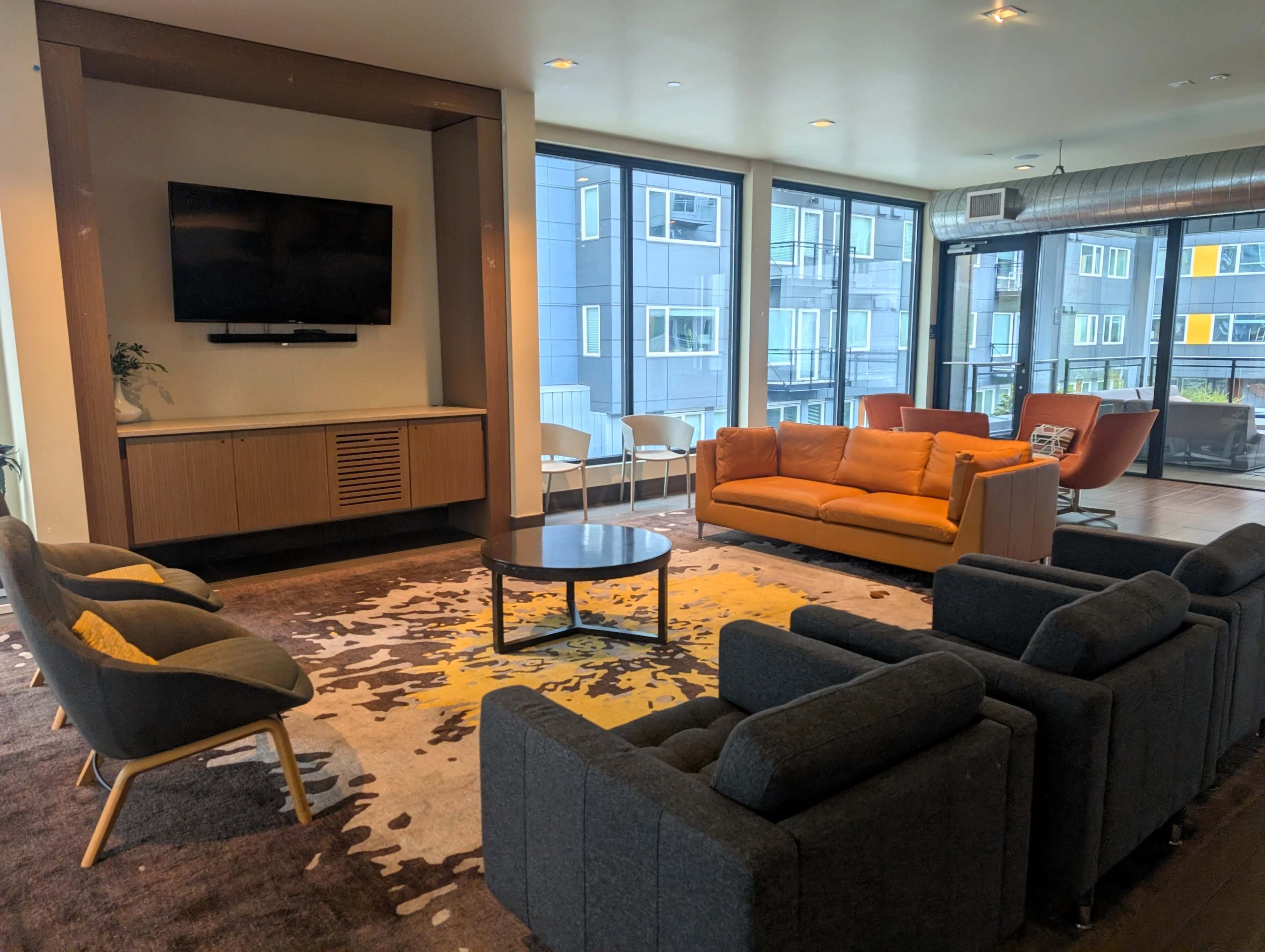 The image shows a modern lounge area with a television mounted on the wall, an orange sofa, gray armchairs, and large windows displaying an urban view.