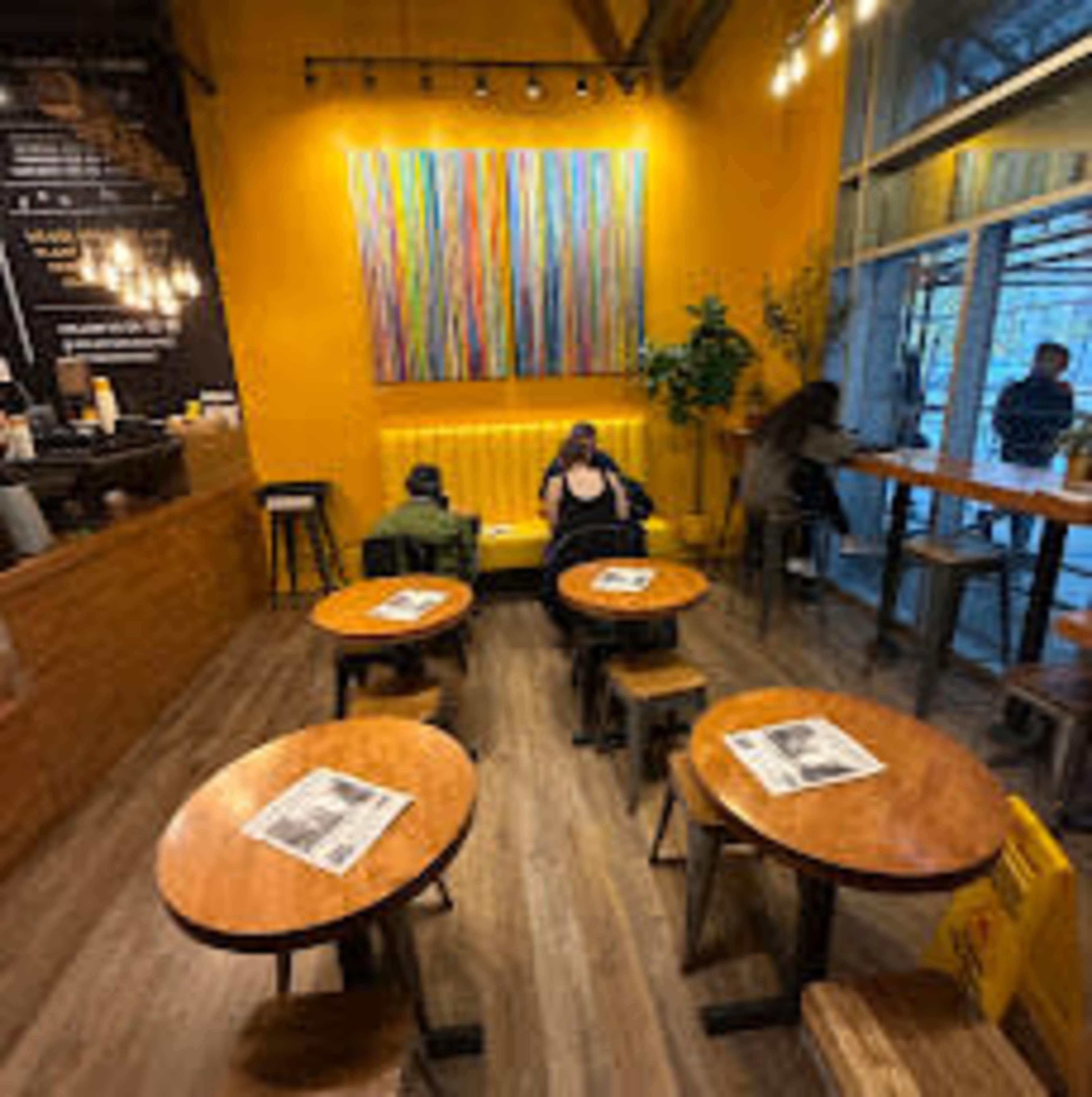 The image shows a cozy café interior with wooden tables and stools, yellow walls, and colorful artwork hanging above a seating area.