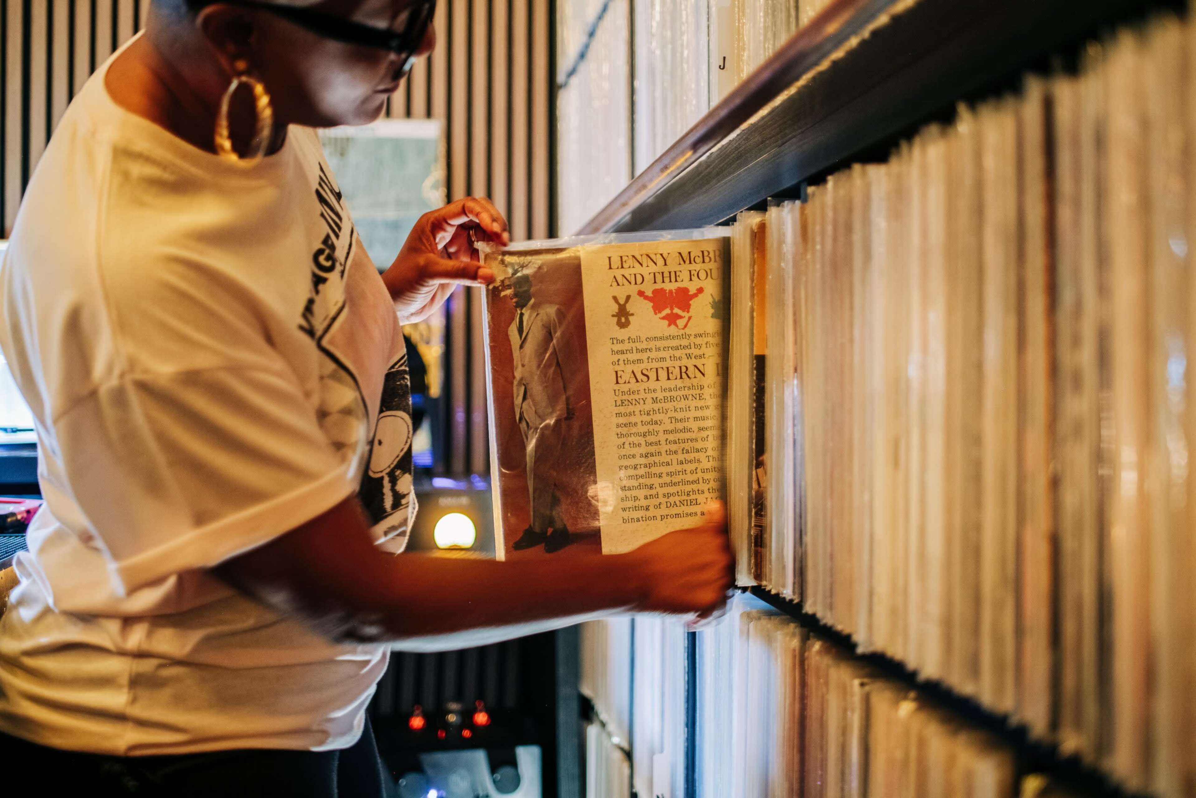 A person with a short hairstyle is pulling out a vinyl record from a wall of neatly organized records.