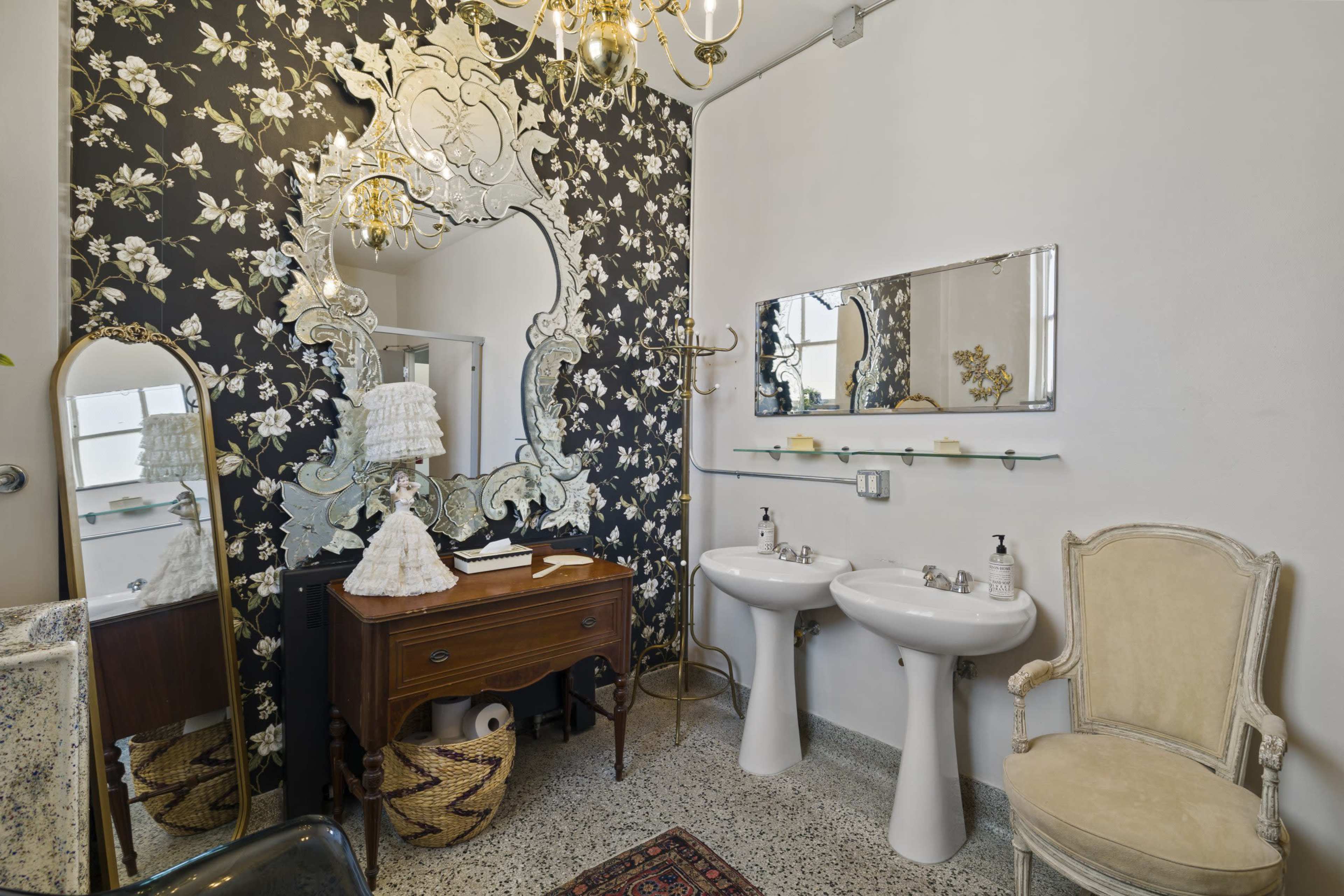 A stylish bathroom features a floral wallpaper, double sinks, a large decorative mirror, and elegant furnishings including a vintage chair and a wooden vanity.
