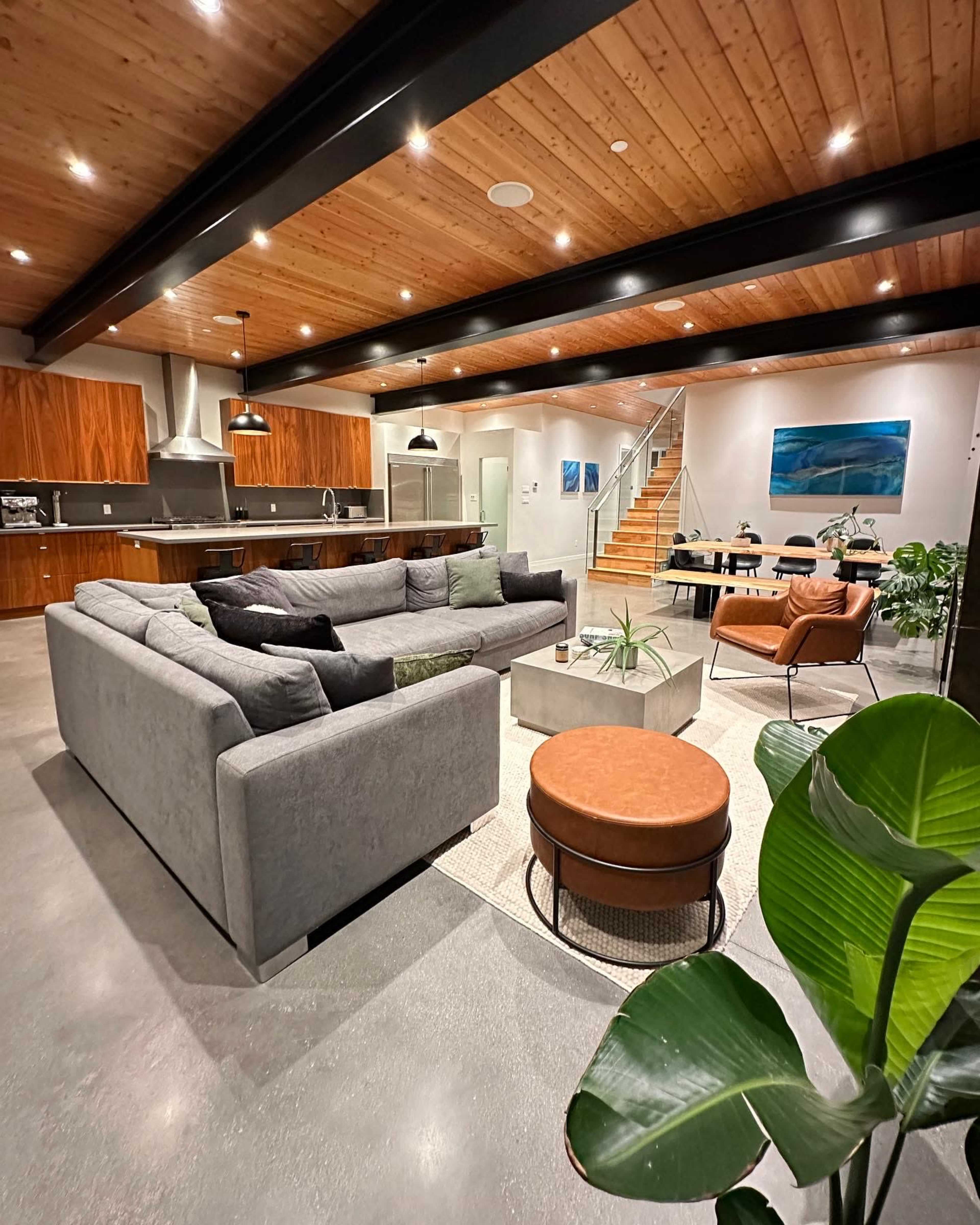 A modern open-concept living space with a large grey sectional sofa, a circular orange ottoman, a dining area, and a kitchen with wooden cabinetry and stainless steel appliances.