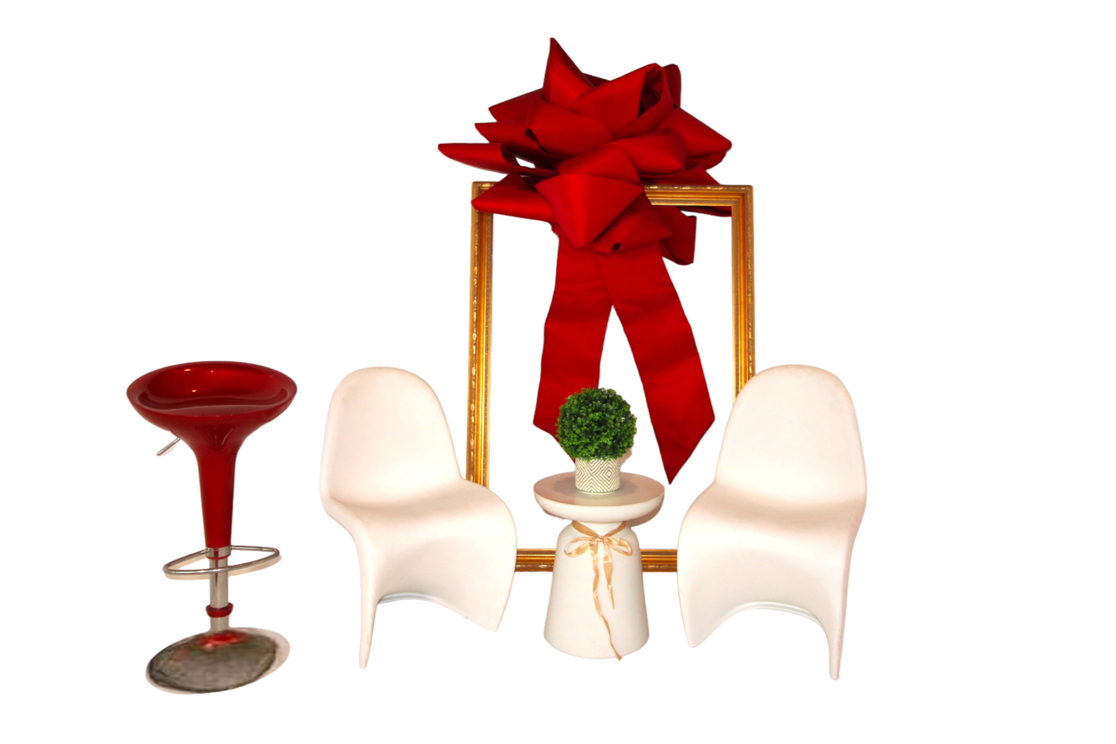 Two white chairs flanking a small table with a potted plant, accompanied by a red bar stool and a large gift-wrapped frame adorned with a red bow at the top.