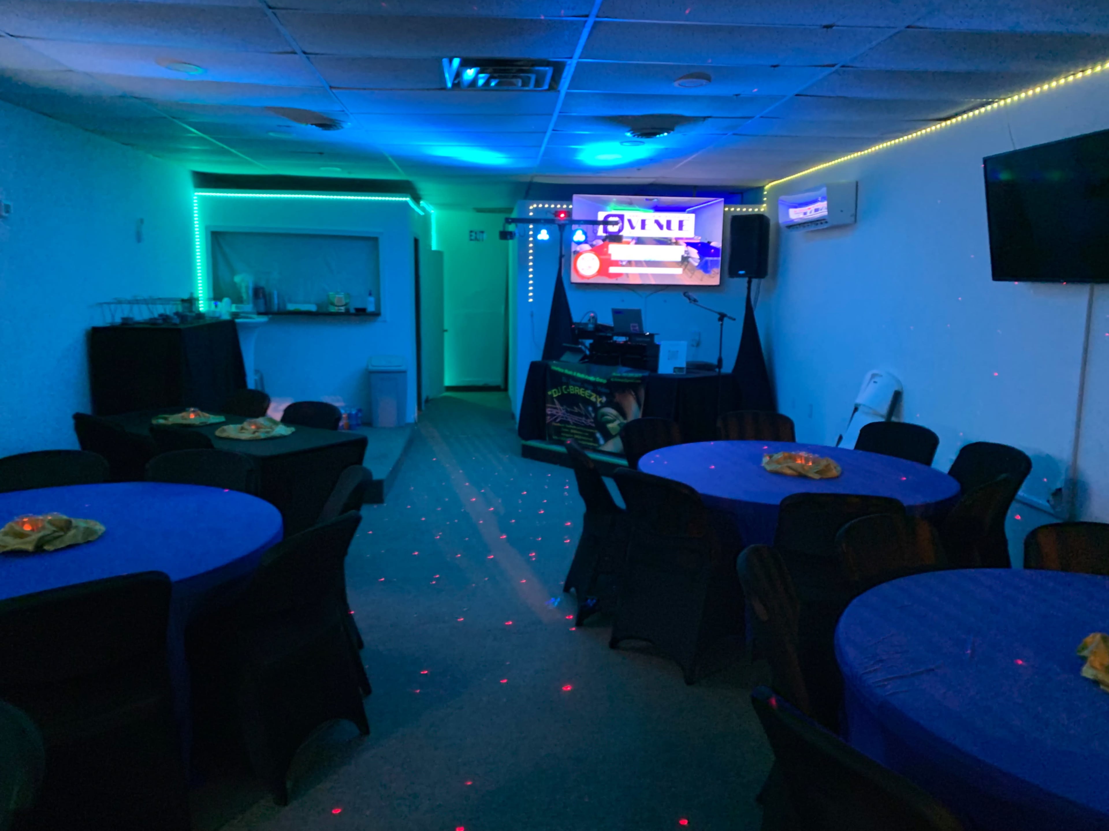 The image shows a dimly lit party room with blue walls, decorated tables covered in blue cloth, a DJ setup in the corner, and colorful lights illuminating the space.