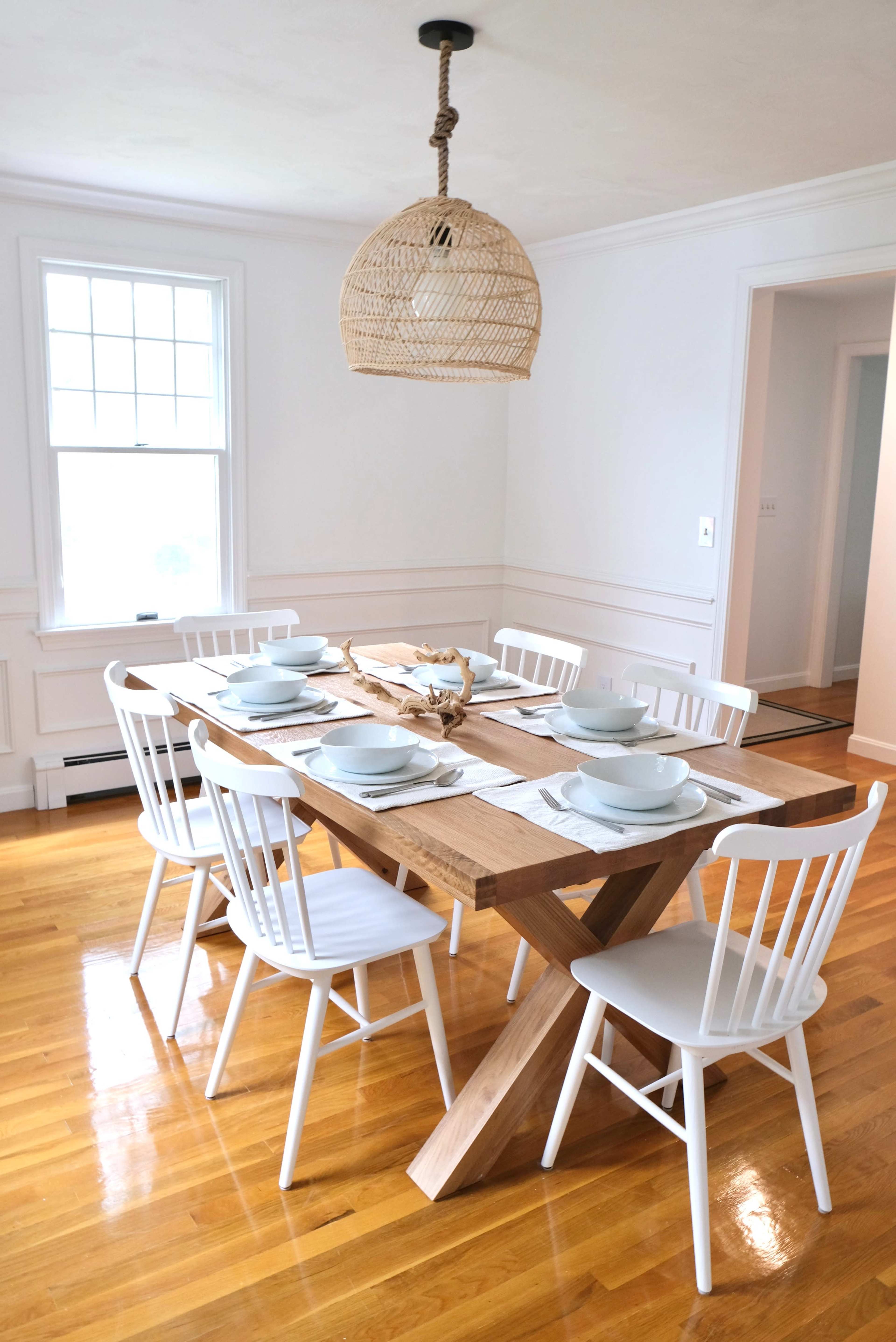 A modern dining room features a large wooden table with eight white chairs, set with blue plates and a natural centerpiece, under a woven pendant light.