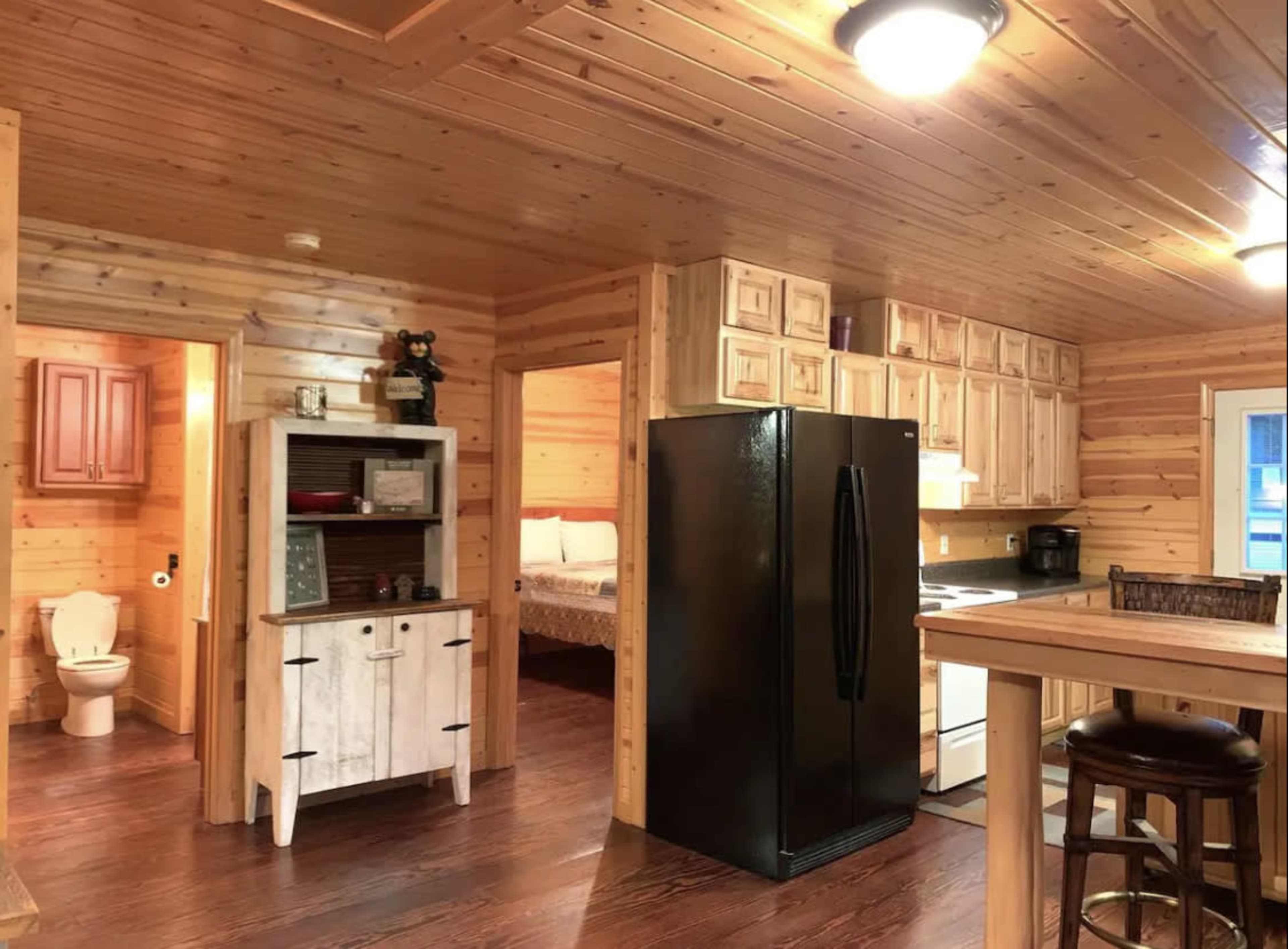 The image shows a wooden interior of a cabin kitchen and living area featuring a black refrigerator, a wooden table, and a small bathroom nook.