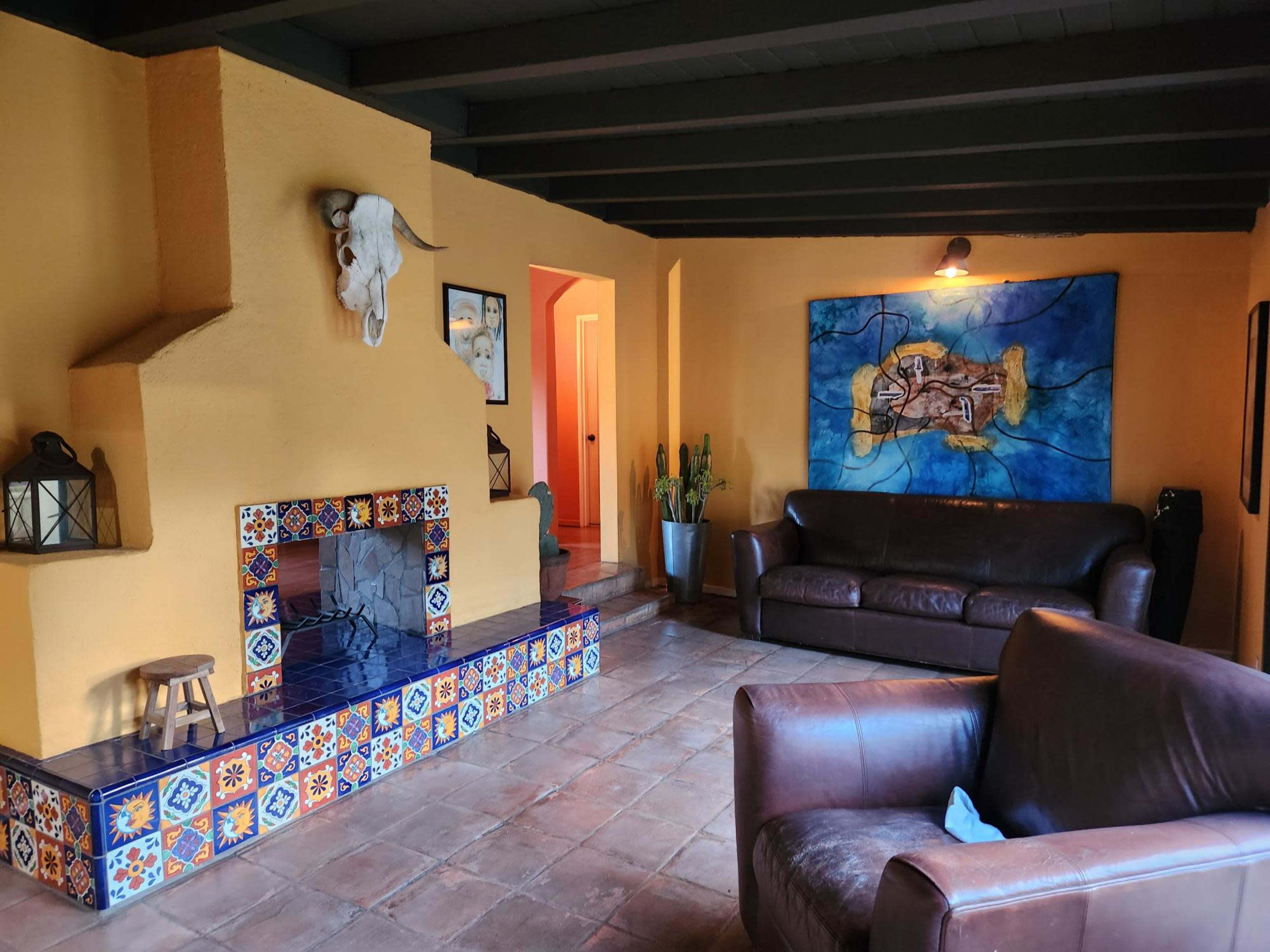 The room features a colorful tiled fireplace, two leather sofas, and a large wall painting, with warm yellow walls and wooden beams overhead.
