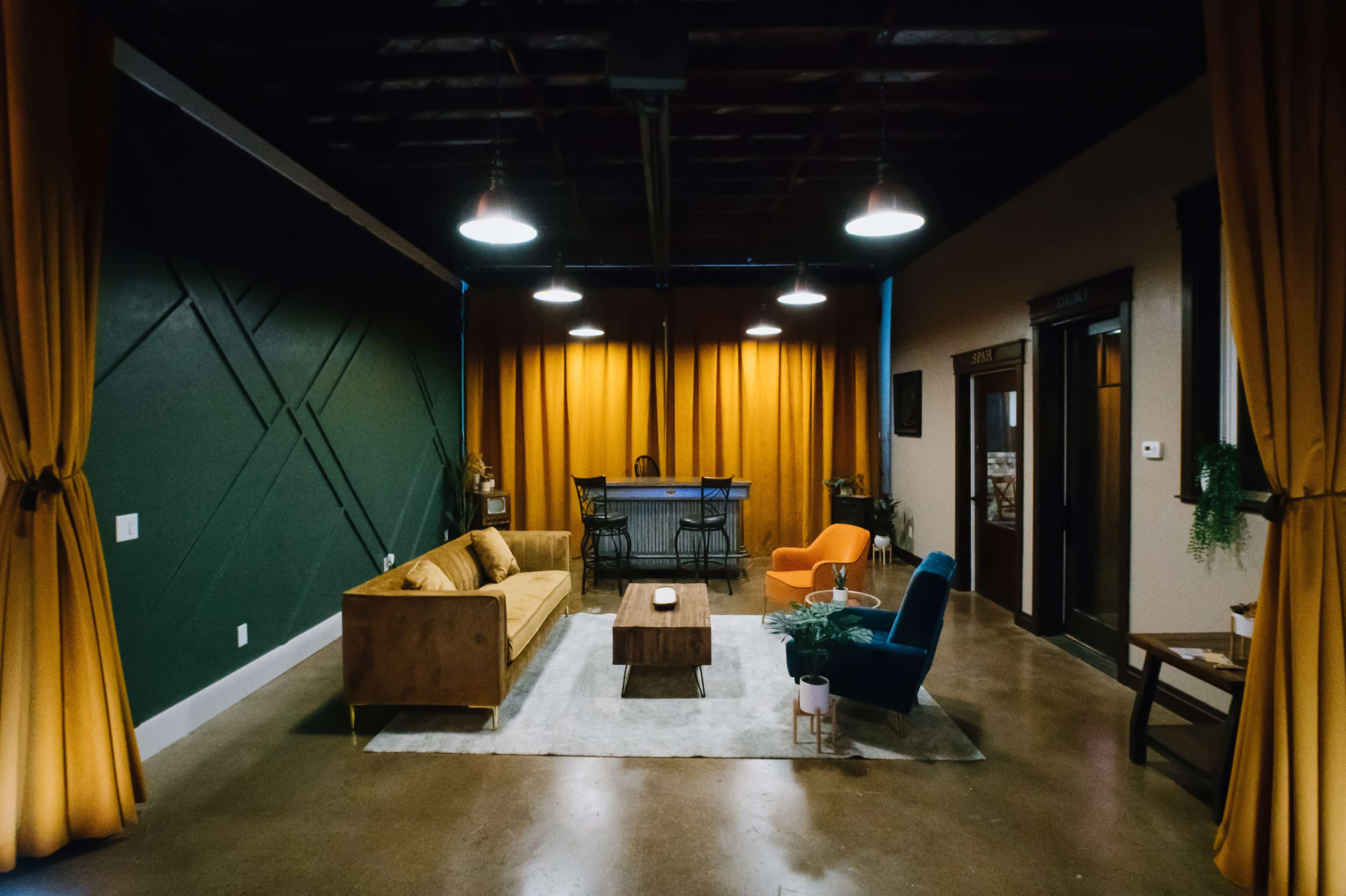 A modern lounge area with a green wall, yellow curtains, a wooden coffee table, and a mix of orange and blue chairs.