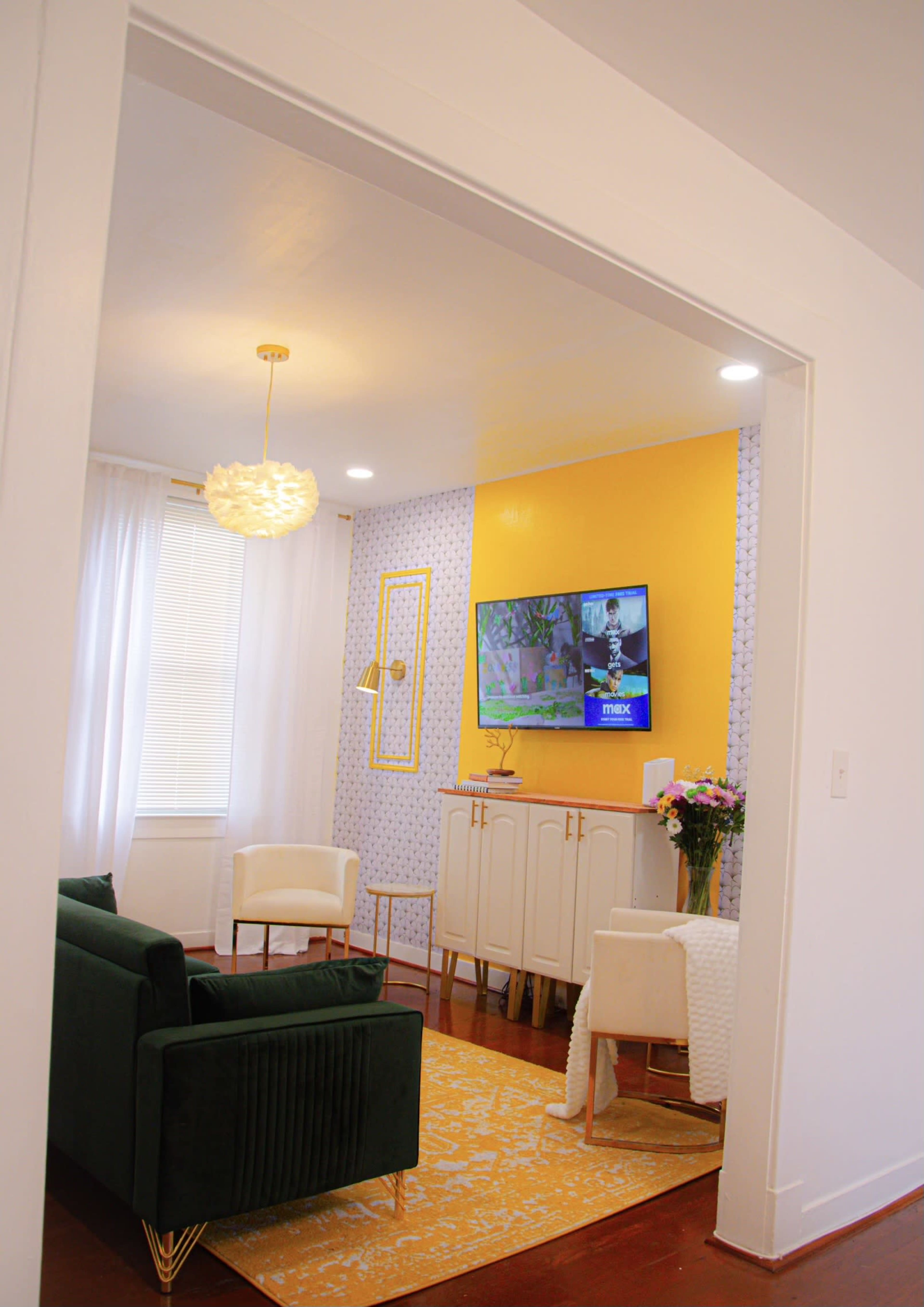 A modern living room is visible through an arched doorway, featuring a green sofa, a white armchair, a yellow accent wall with a TV mounted on it, and a decorative rug.