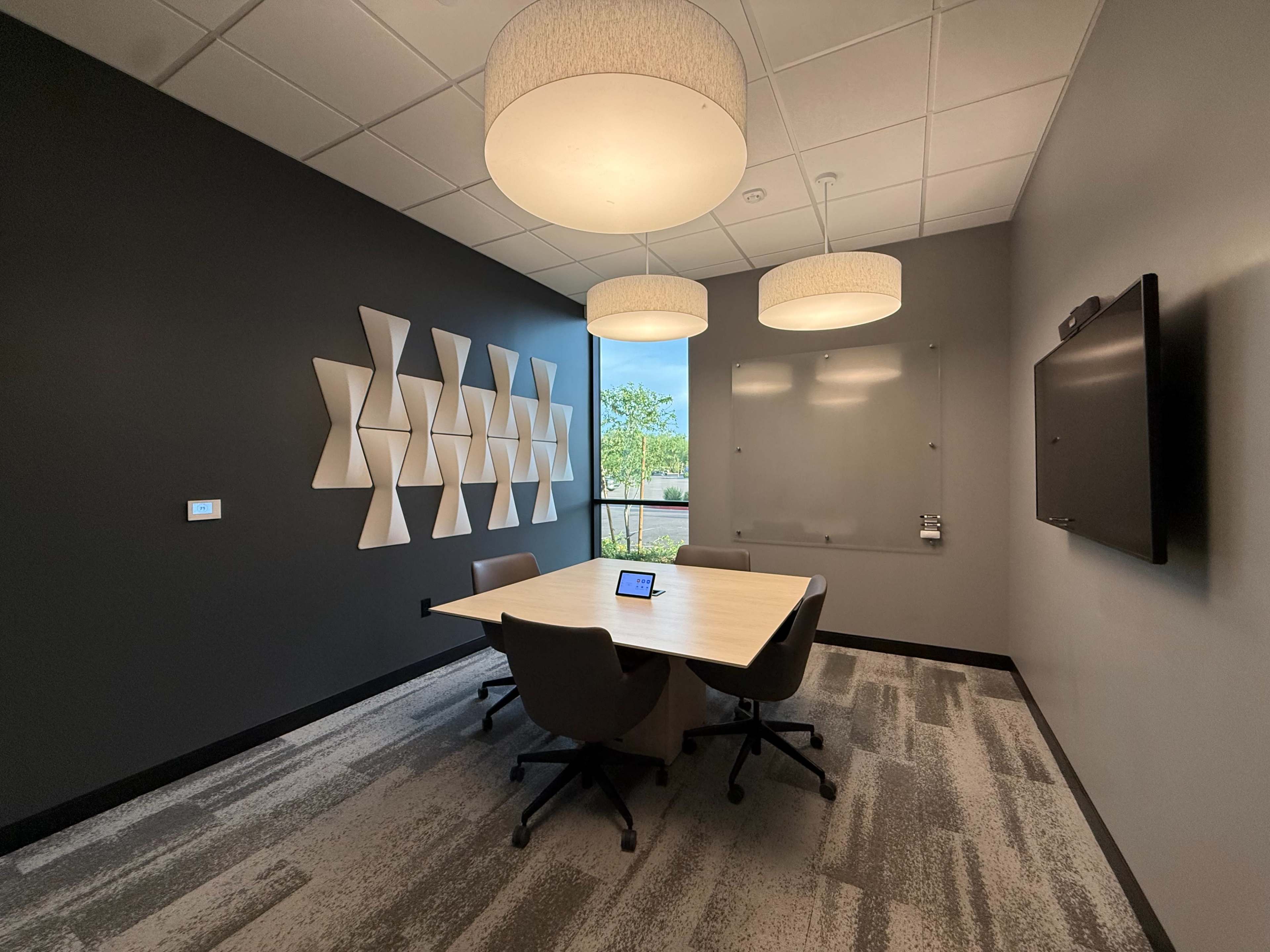 The image shows a modern conference room with a rectangular table, four chairs, and three overhead light fixtures, complemented by a wall of geometric shapes and a large display screen.