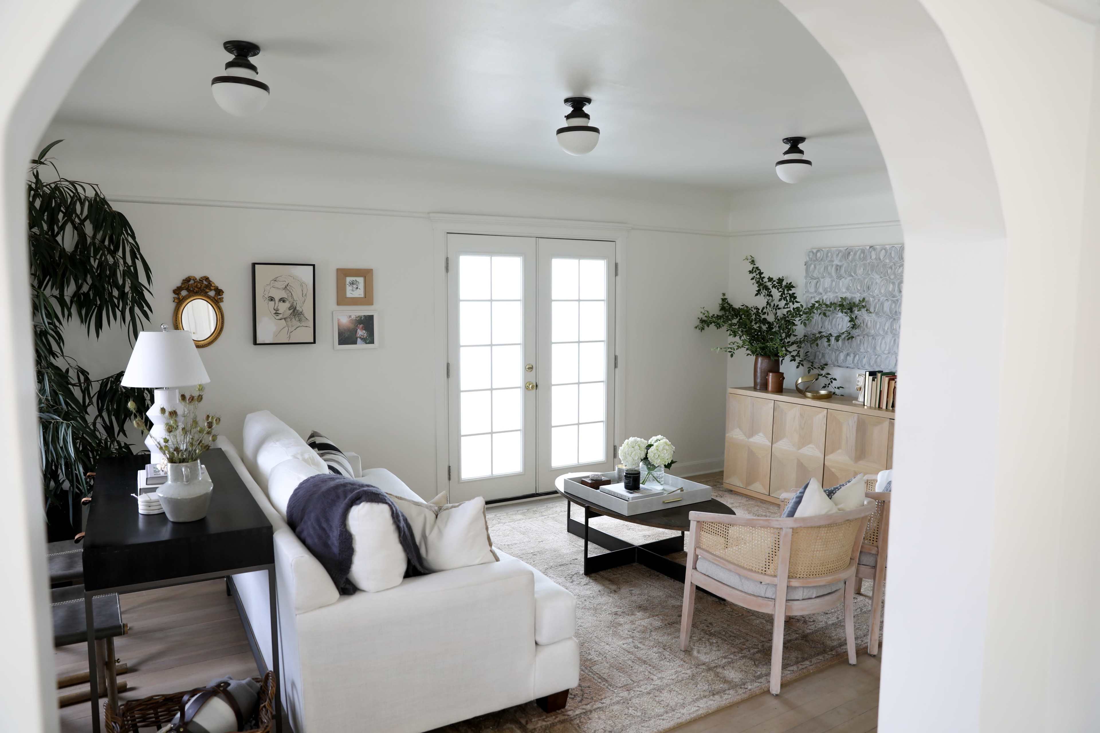A bright living room features a white sofa, a wooden coffee table, and double French doors opening to an outdoor space.