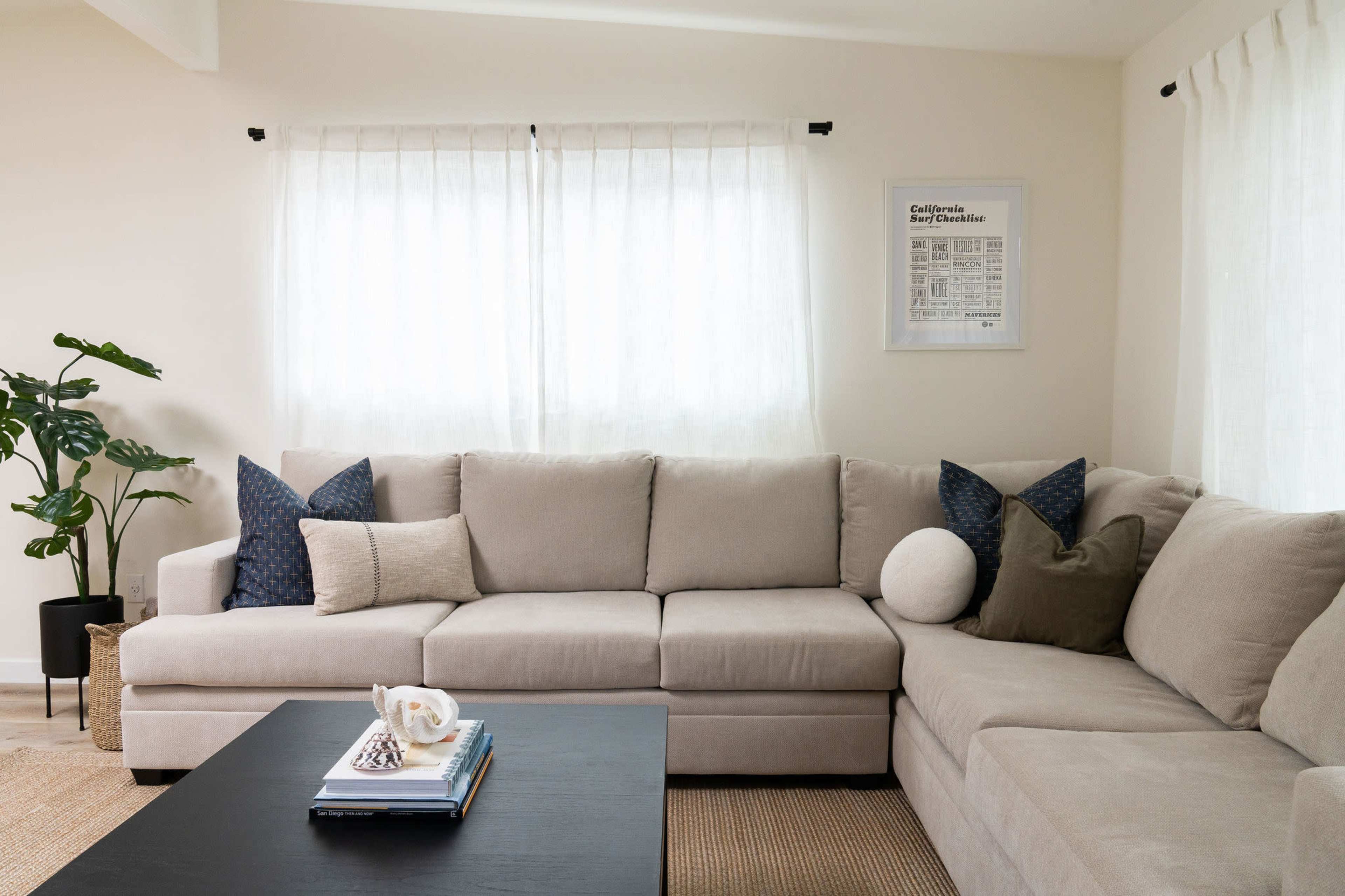 A light-colored sectional sofa with decorative pillows is positioned around a black coffee table in a bright living room featuring two windows with sheer curtains and a potted plant.