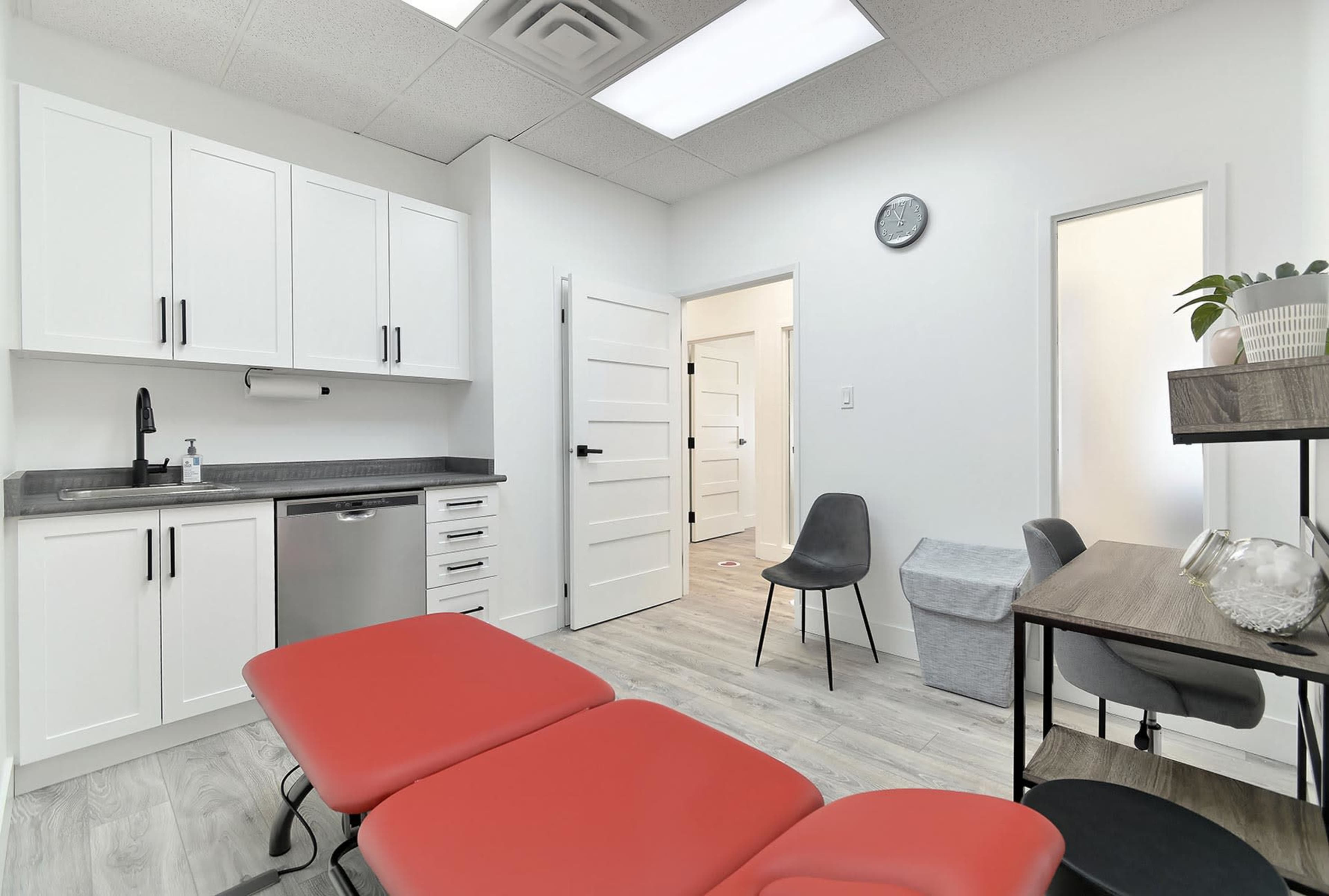A clean, modern medical examination room featuring red examination tables, cabinets, and minimalist furniture.