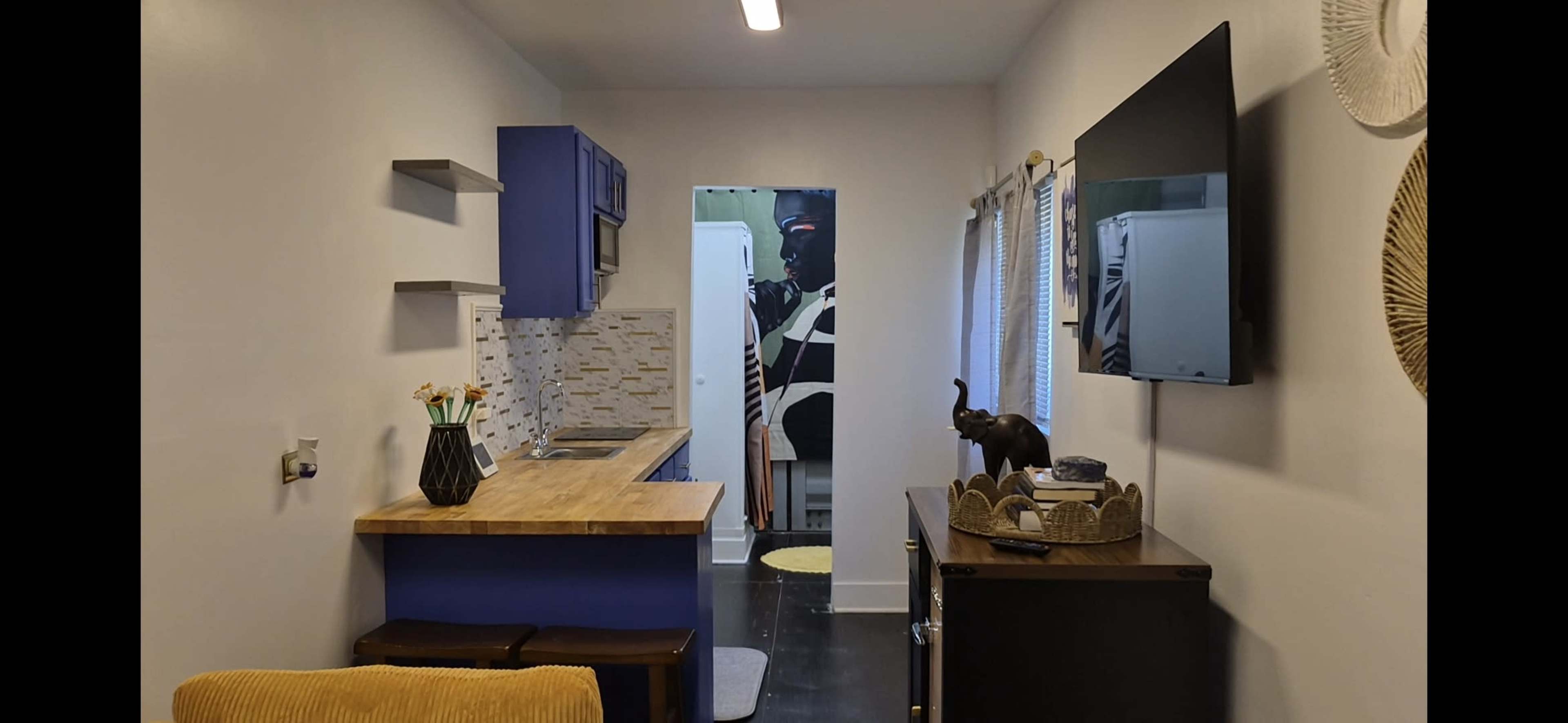 The image shows a compact kitchen area with blue cabinets, a wooden countertop, and a television mounted on the wall opposite a small dining table and chairs.