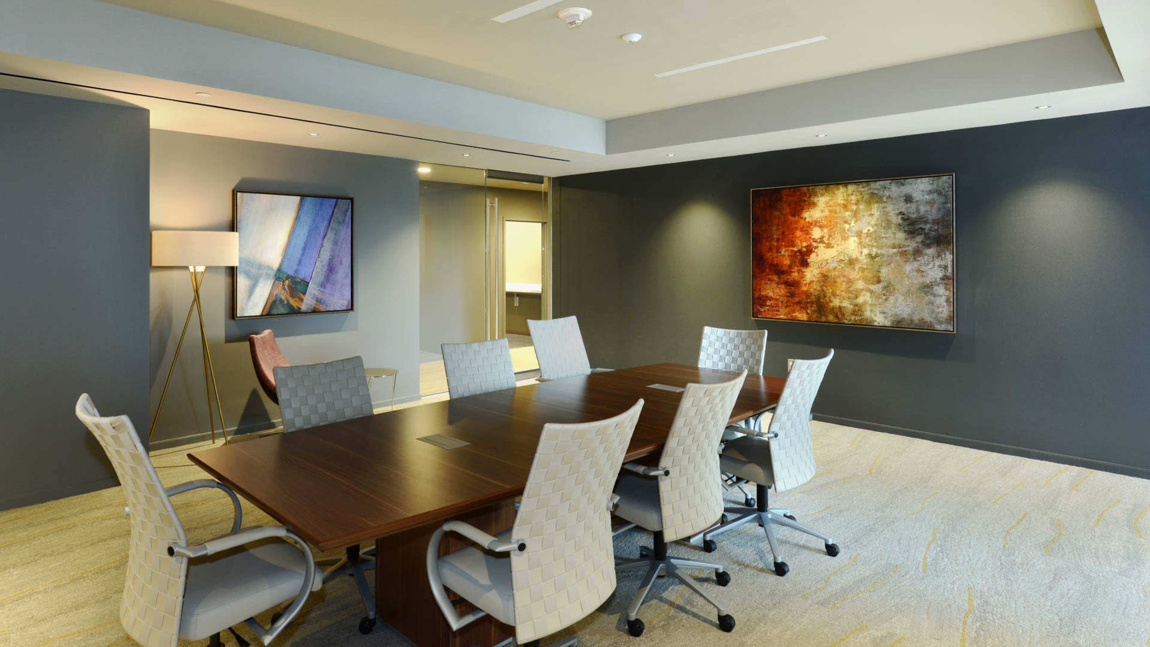 A modern conference room features a large wooden table surrounded by eight ergonomic chairs, with abstract artwork on the walls and a lamp in one corner.