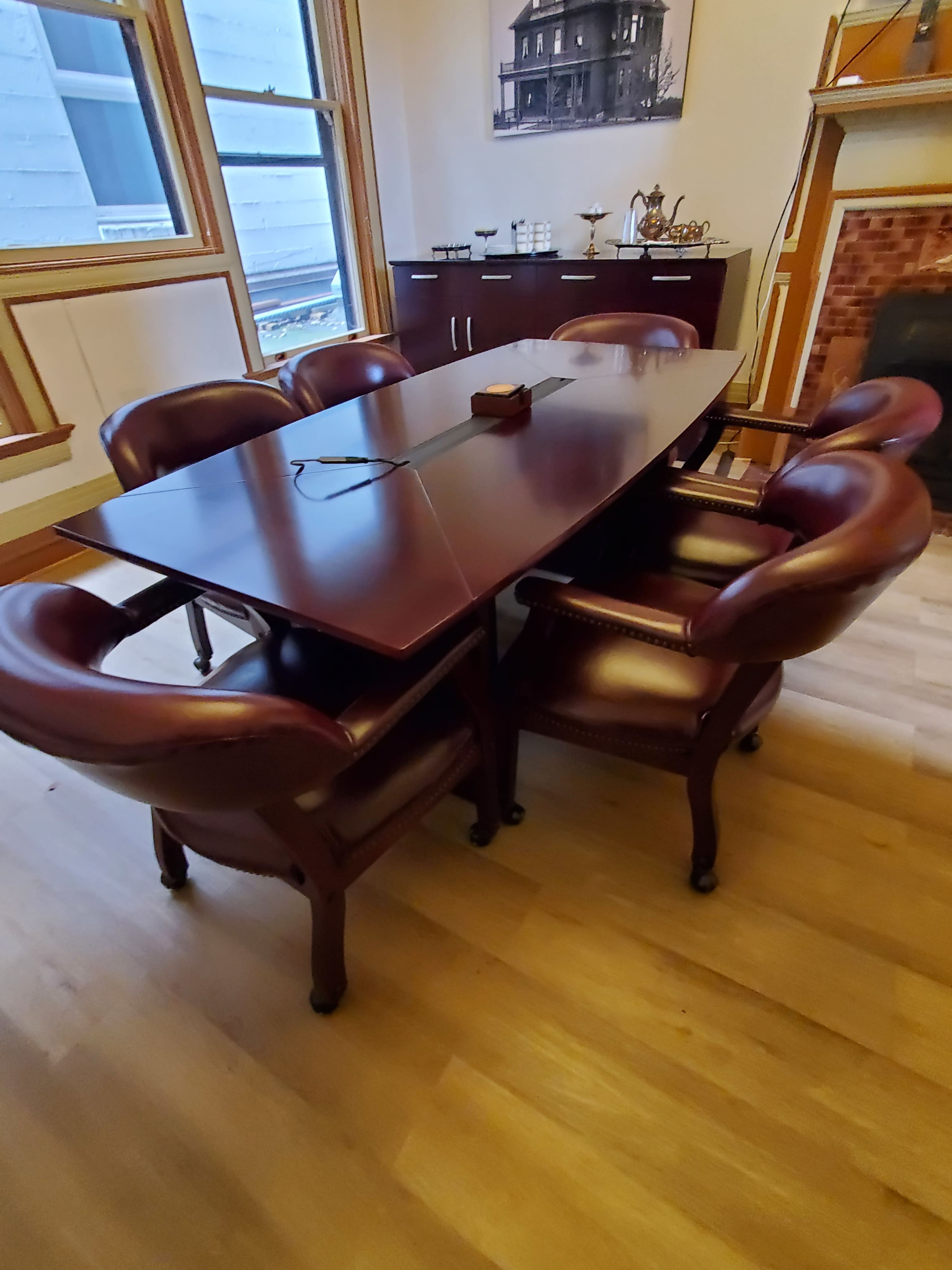 A large wooden table surrounded by six upholstered chairs is set in a room with wooden flooring and a fireplace.