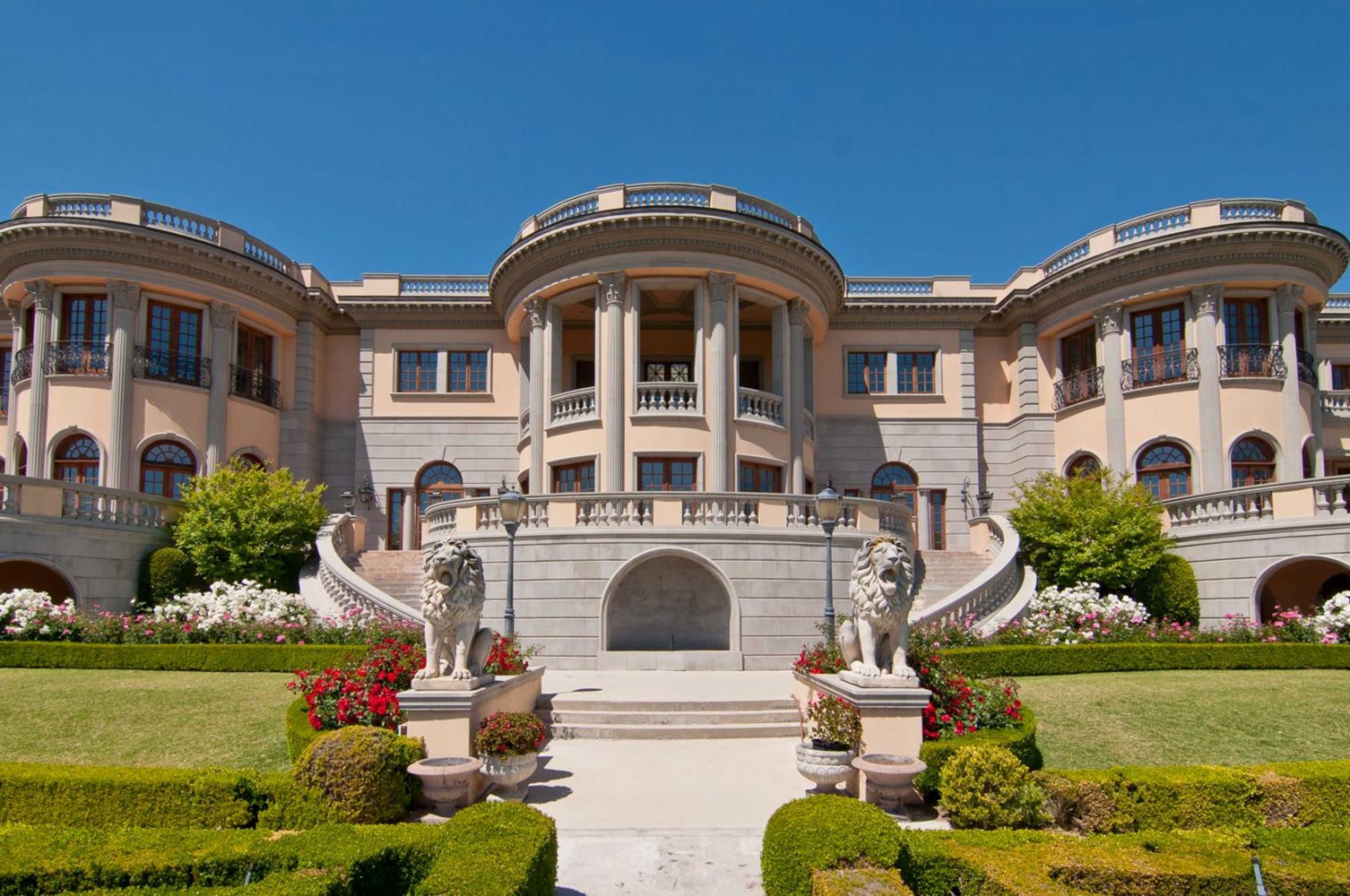 A large, ornate mansion with a symmetrical facade, featuring a grand entrance flanked by lion statues and surrounded by meticulously landscaped gardens.