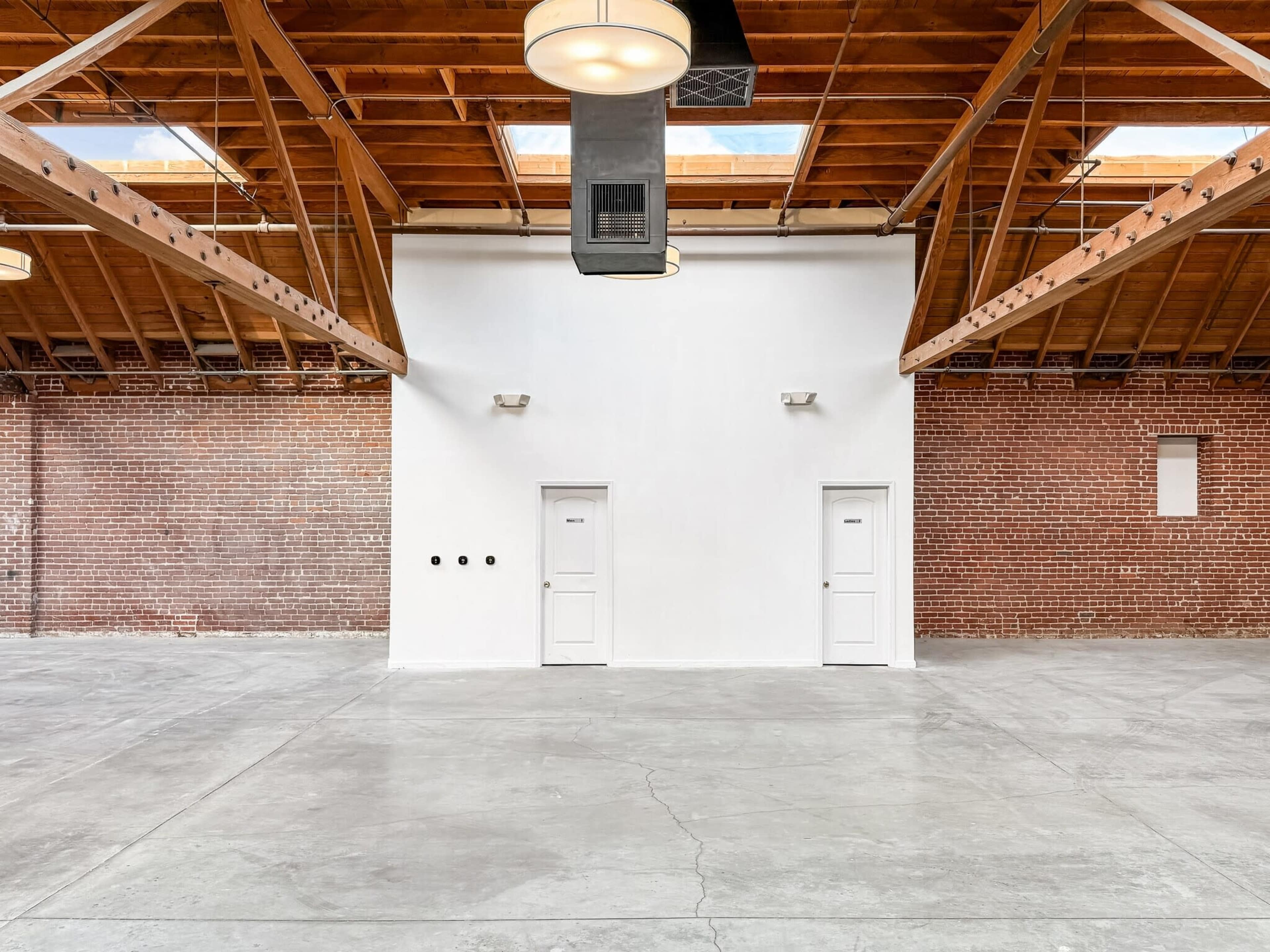 The image shows a spacious, industrial-style interior featuring exposed wooden beams, brick walls, and two white doors on a plain wall.