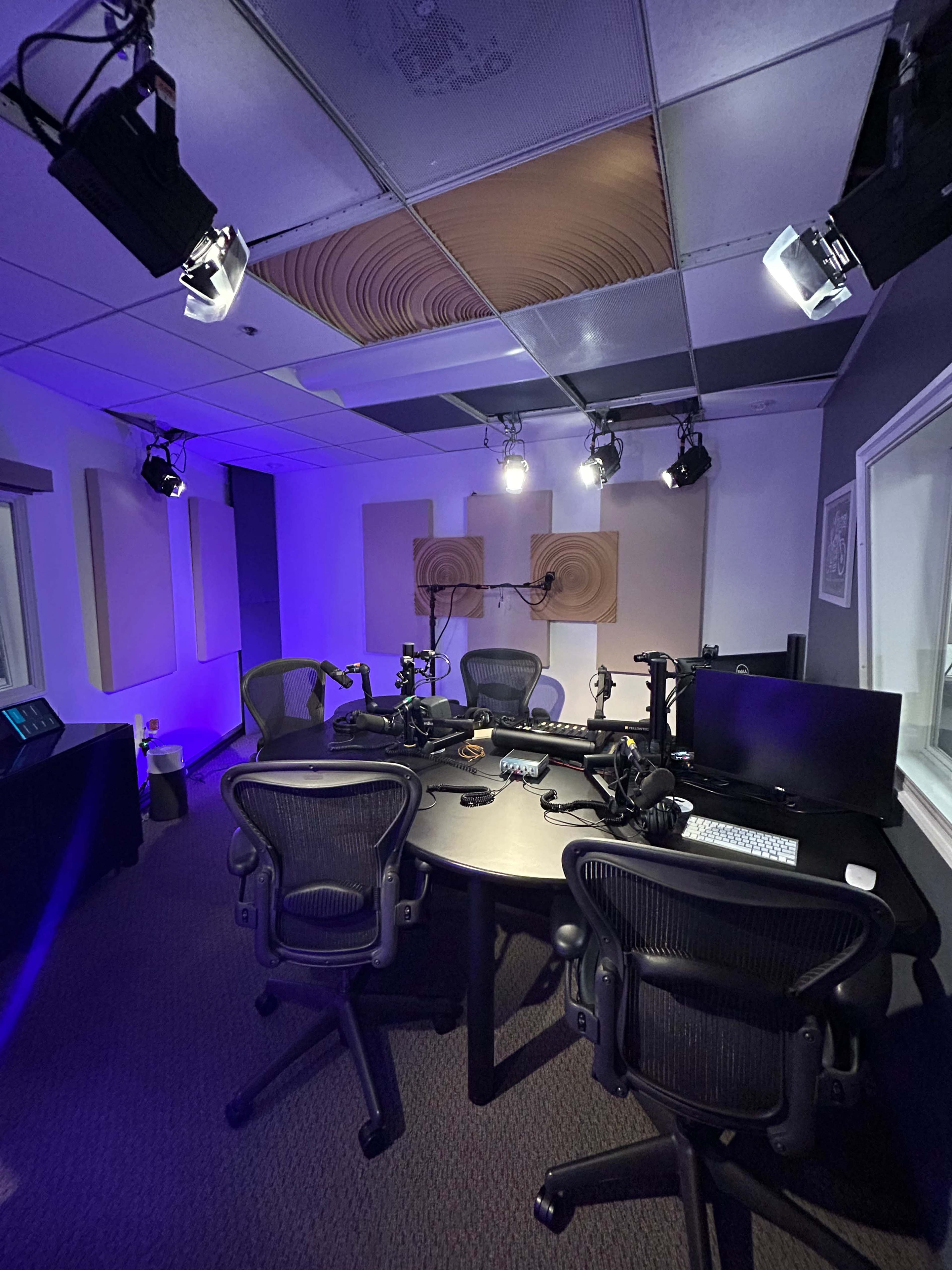 A modern recording studio with several chairs, microphones, and sound equipment arranged around a central black table under blue and white lighting.