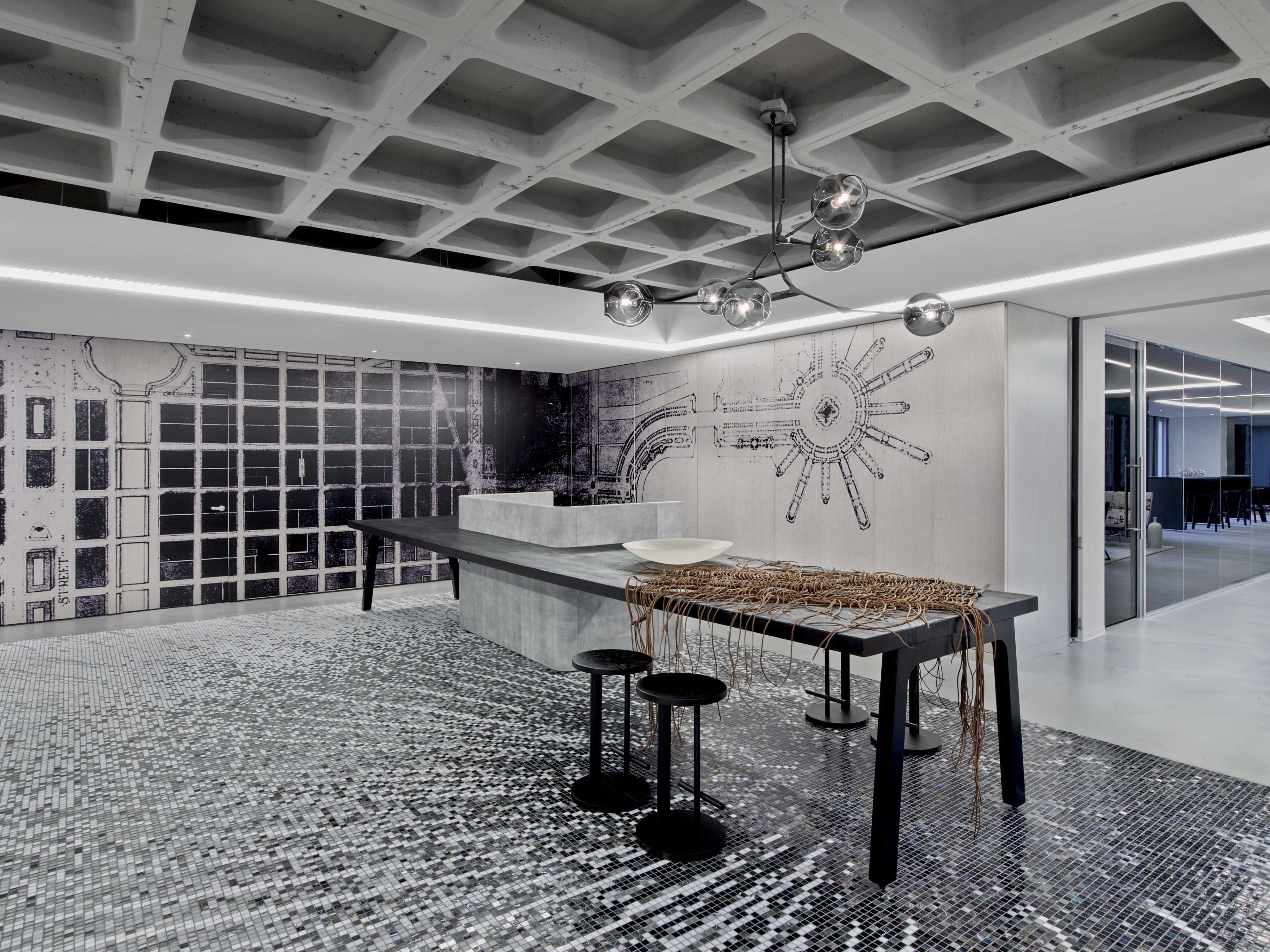 The image shows a modern interior space featuring a patterned floor made of small tiles, a long rectangular kitchen island, a unique light fixture, and graphic wall designs.