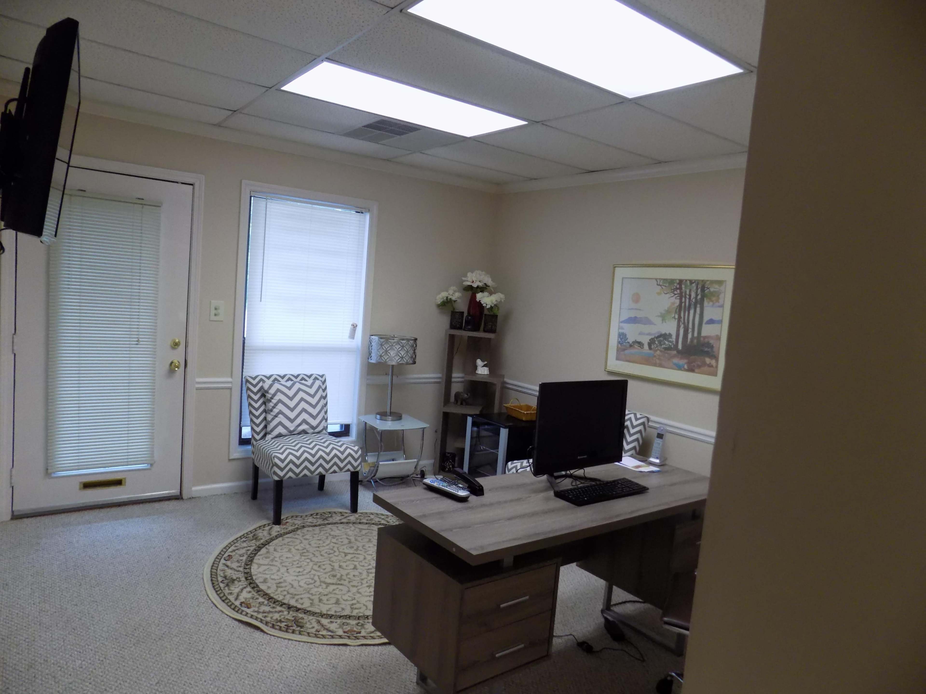 The image shows a small office space with a desk, computer, two chairs, and a decorative rug.