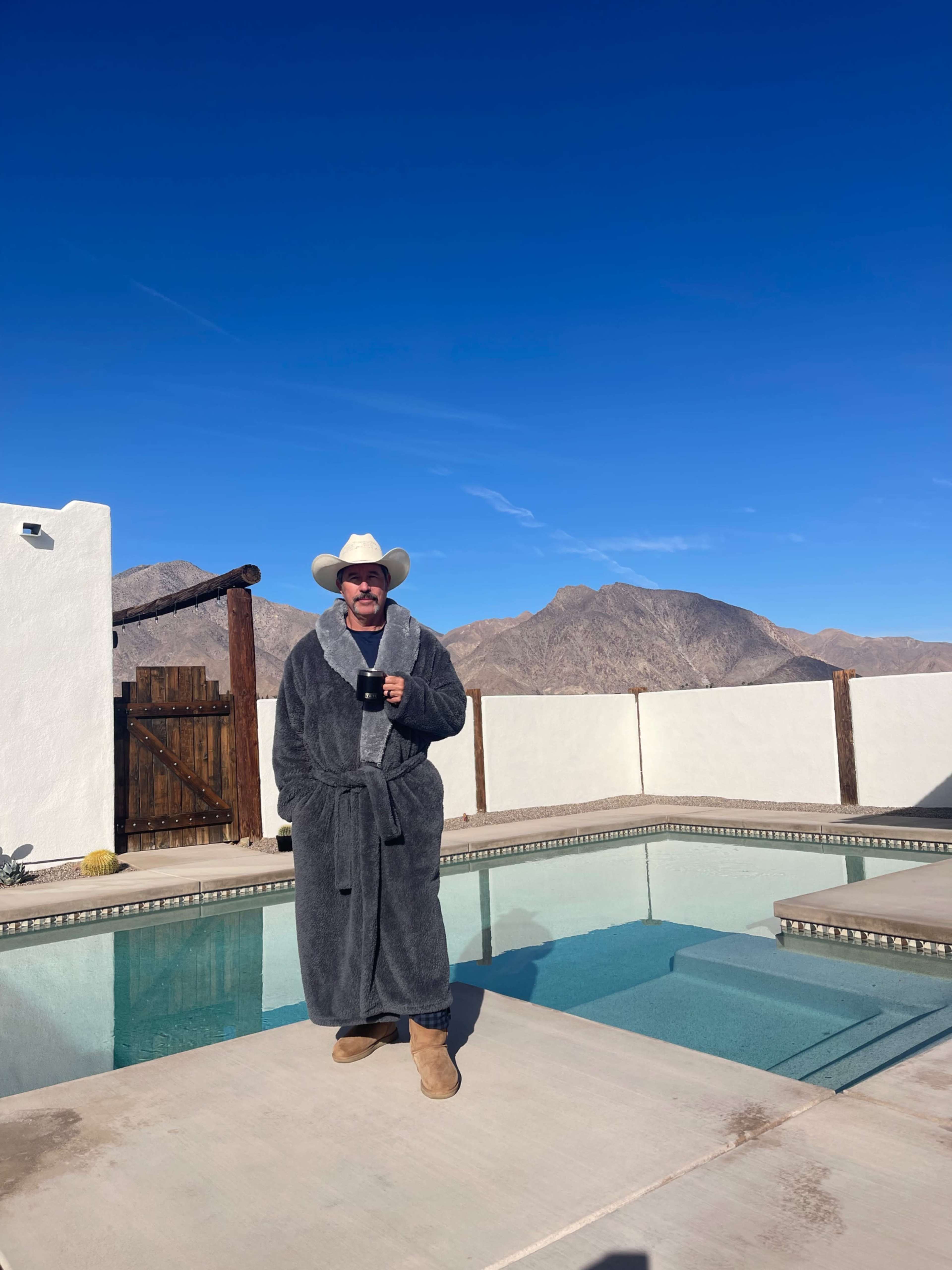 A person in a gray robe and cowboy hat stands by a swimming pool, with mountains visible in the background under a clear blue sky.