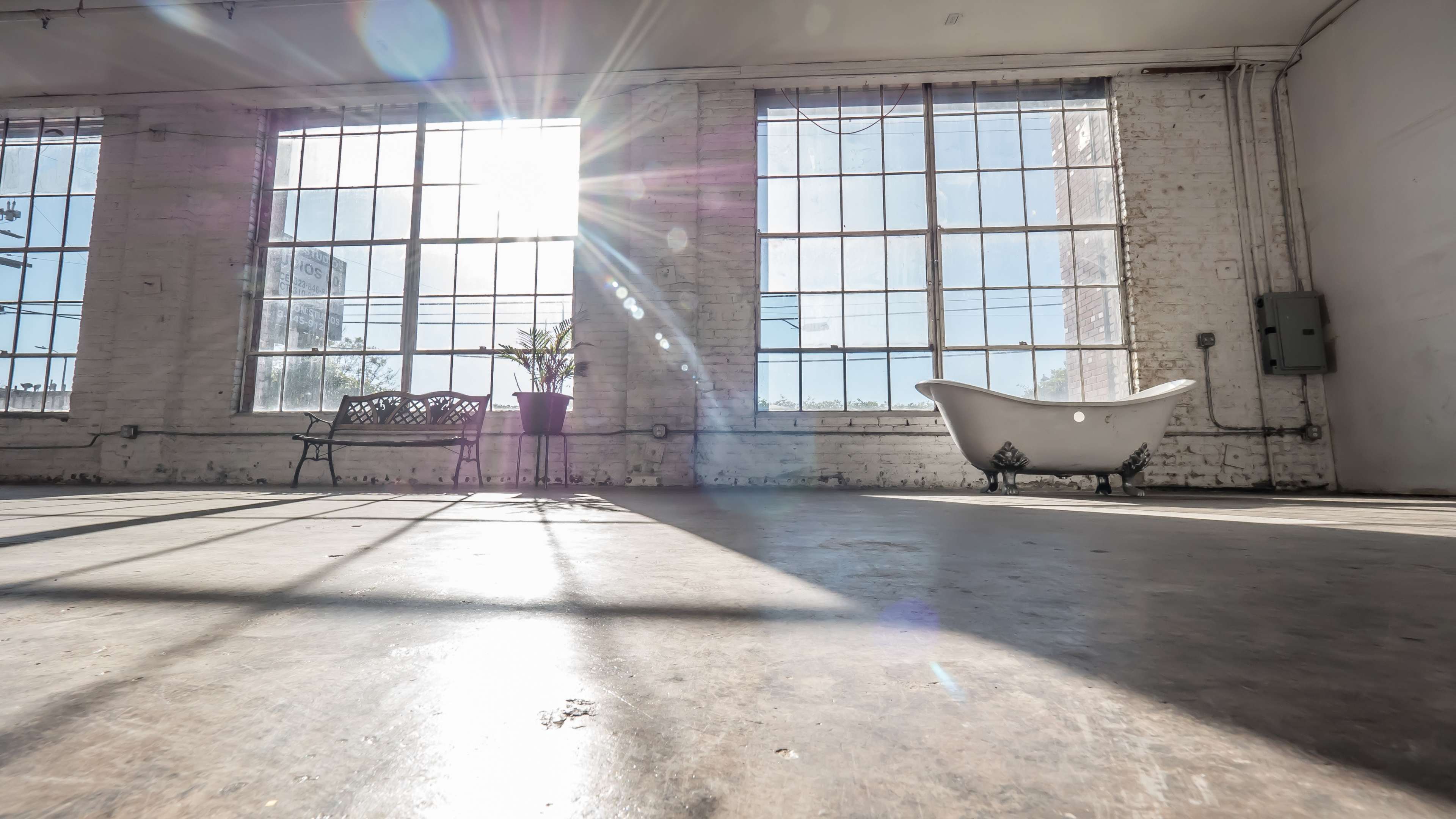 Bright sunlight streams through large windows in a spacious, empty room featuring a vintage bathtub and a bench.