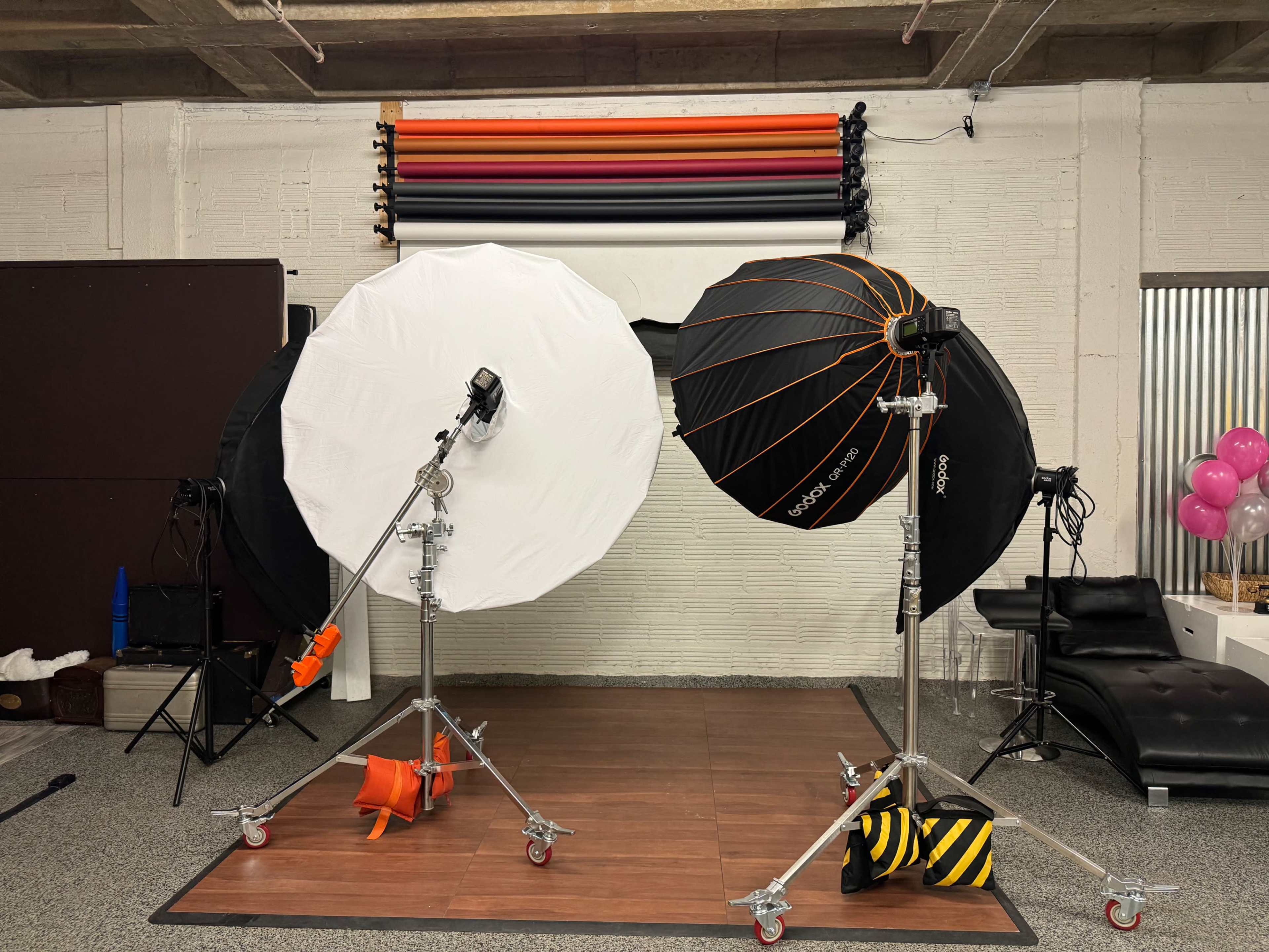 A photography studio setup with two large light modifiers on stands, positioned on a wooden floor against a backdrop of colorful rolled paper.