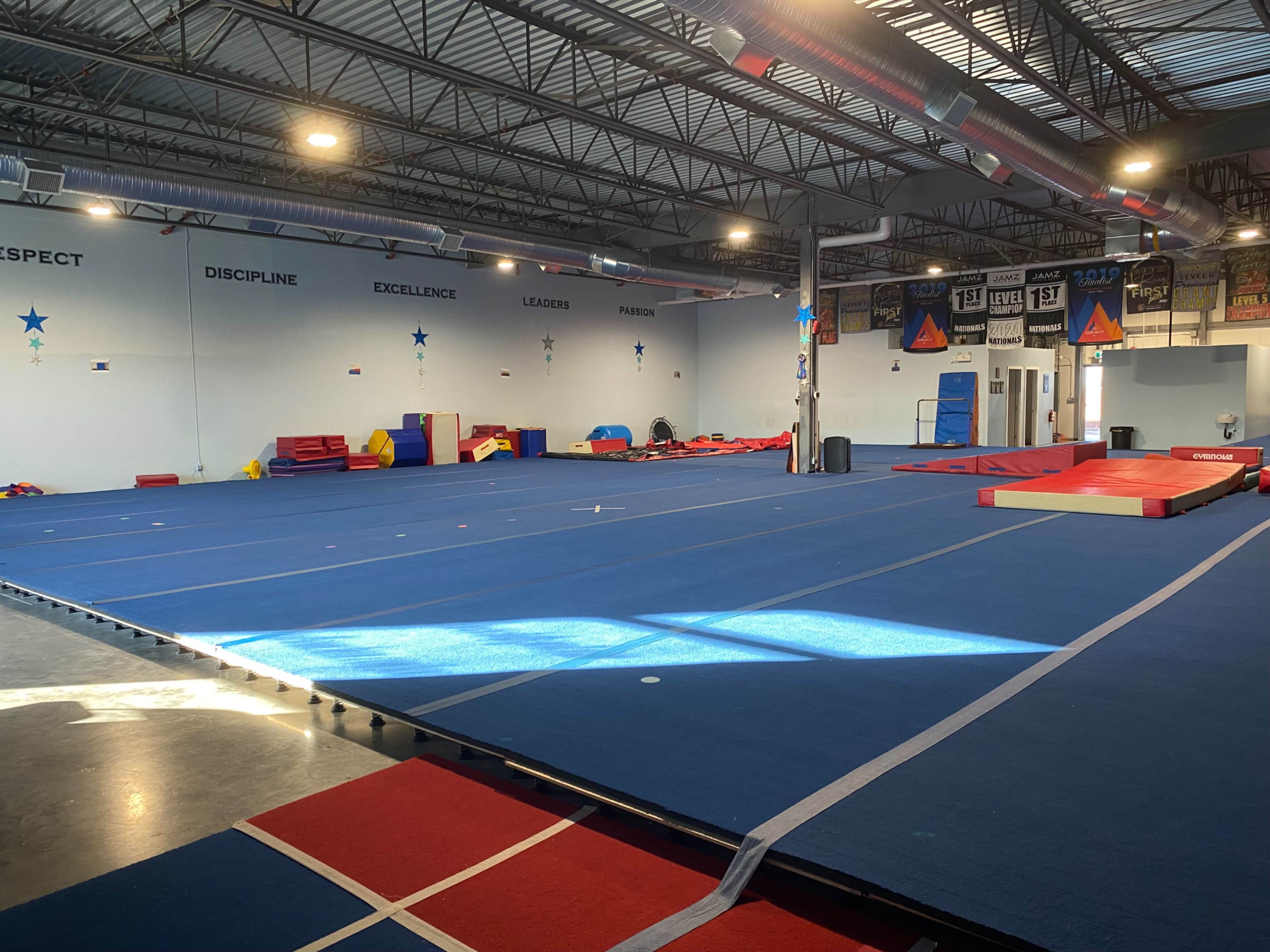 A spacious gymnasium with blue mats, various gymnastic equipment, and motivational banners on the walls.