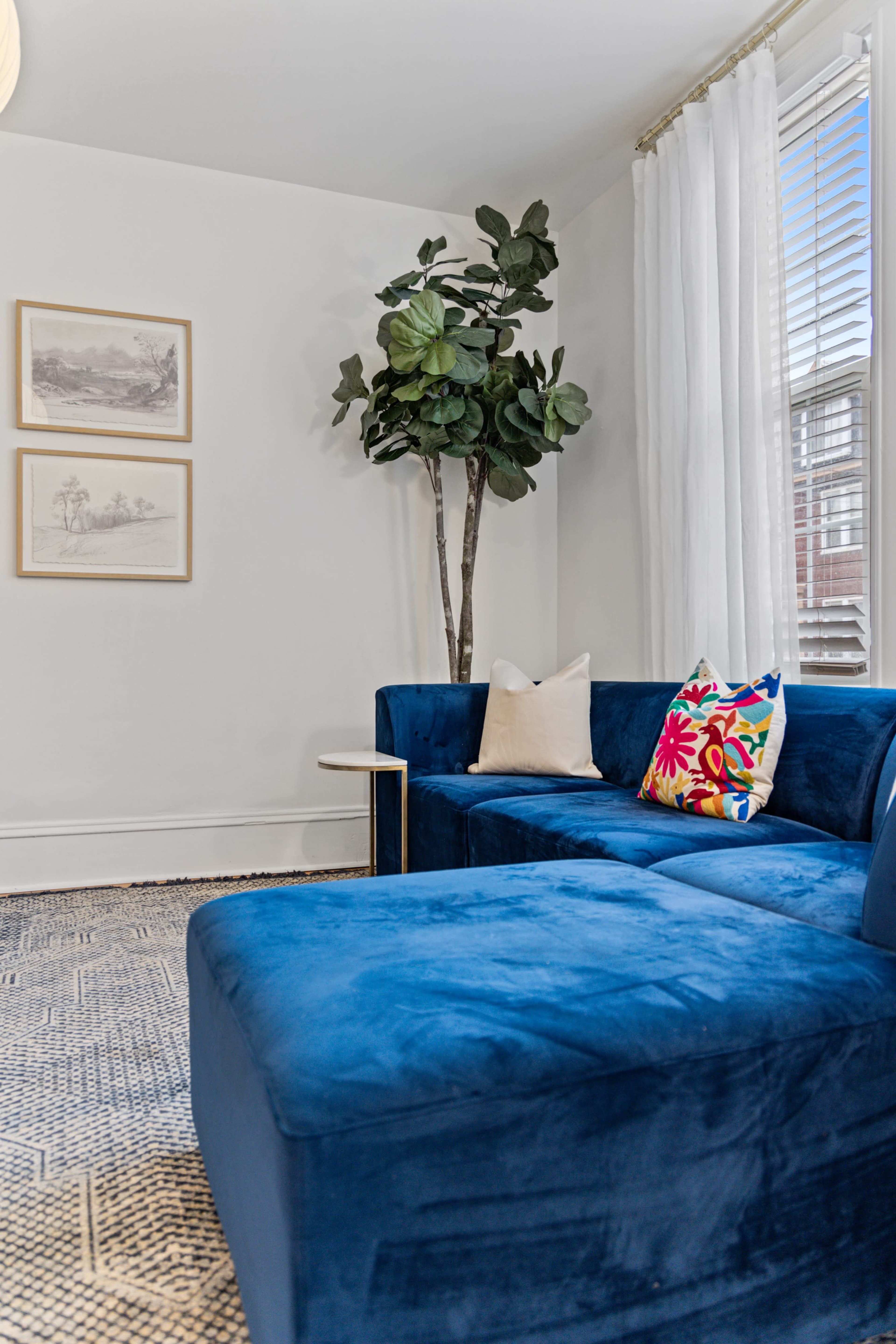 A blue velvet sectional couch with colorful pillows is positioned next to a potted plant and framed artwork on the wall.
