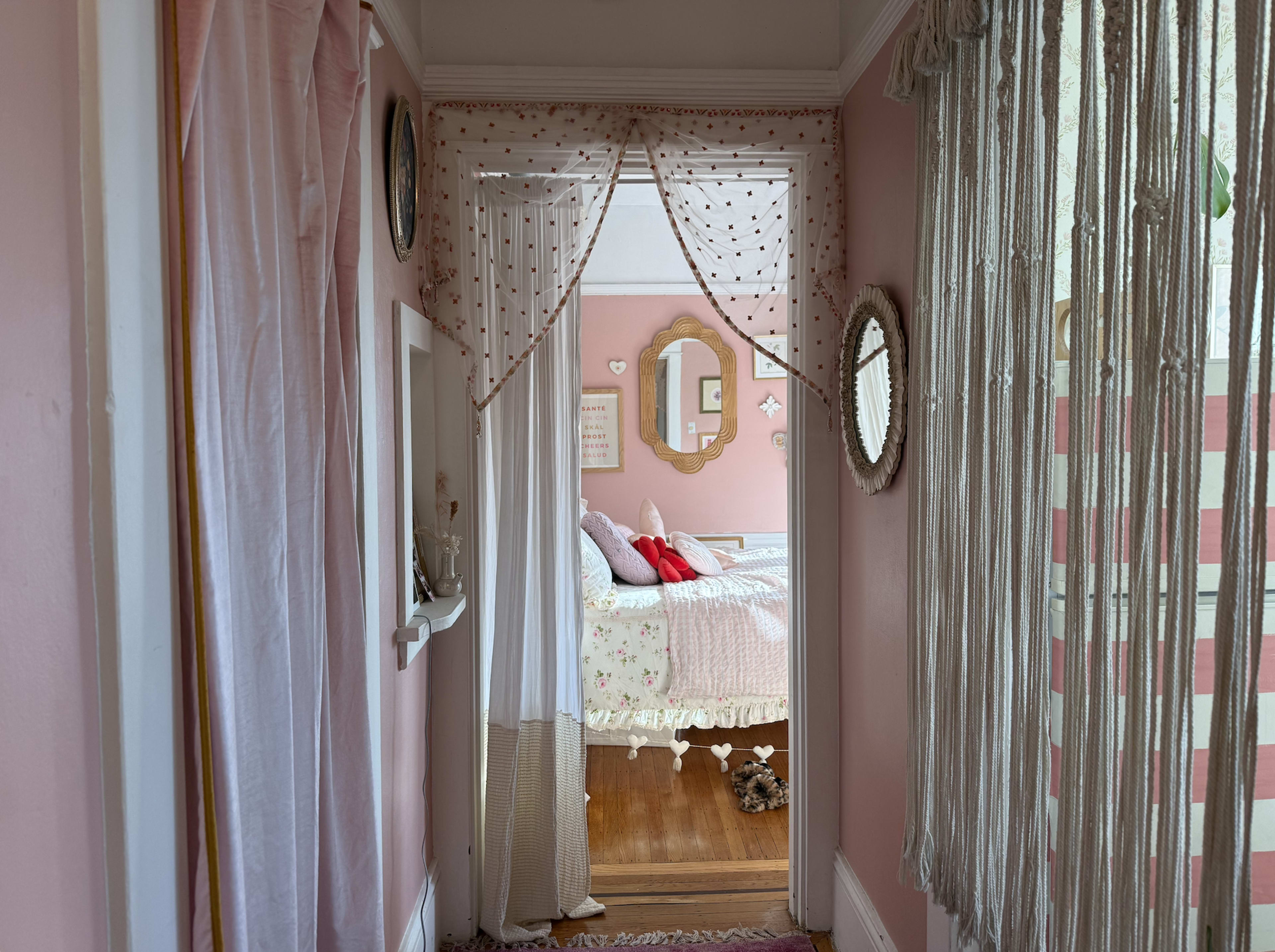 The image shows a pink-walled hallway leading to a bedroom adorned with floral decorations and a bed with red and white pillows.
