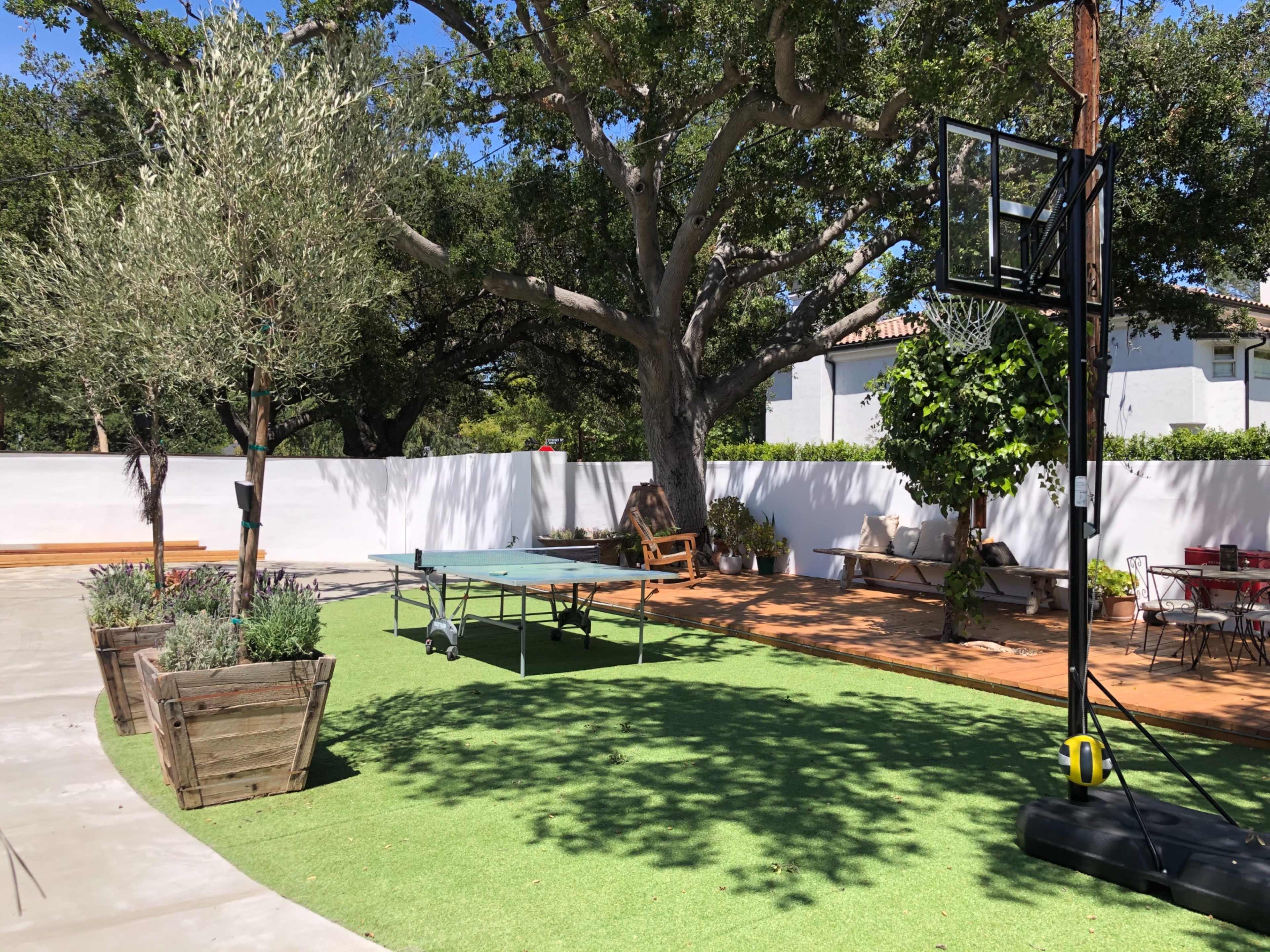 The image shows a backyard space featuring a basketball hoop, a ping pong table, and landscaped areas with trees and planters.