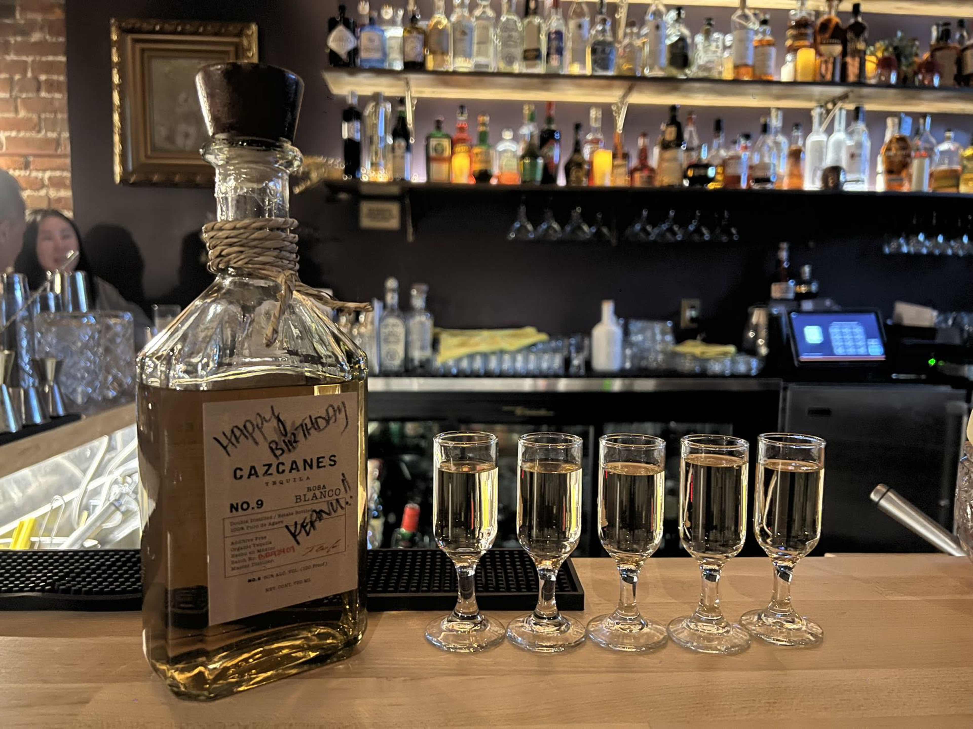 A bottle of Cazcaanes tequila is displayed on a bar with five shot glasses arranged in a row in front of it.