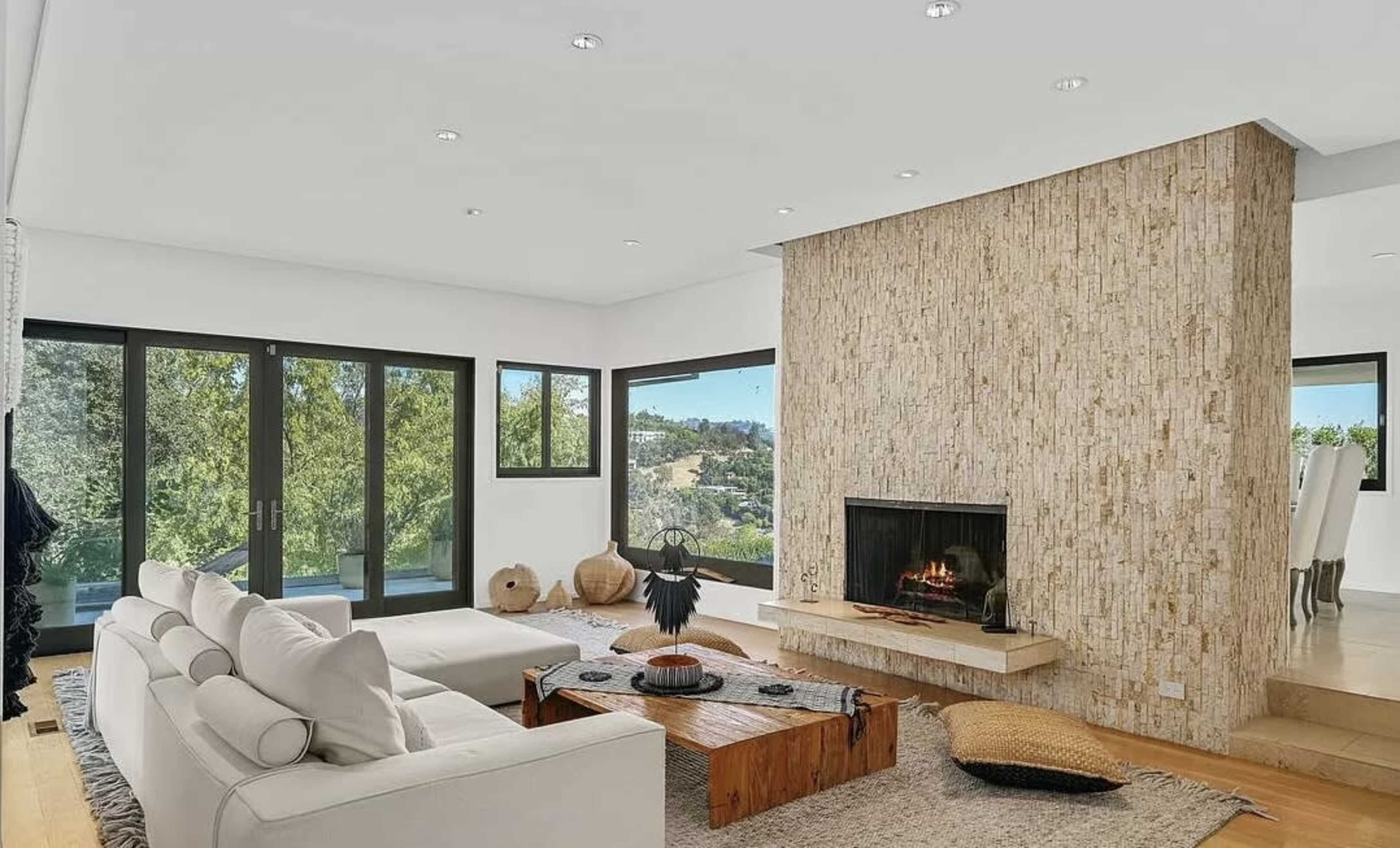 The living room features a large white sofa, a wooden coffee table, and a textured stone fireplace, surrounded by large windows that overlook a green landscape.