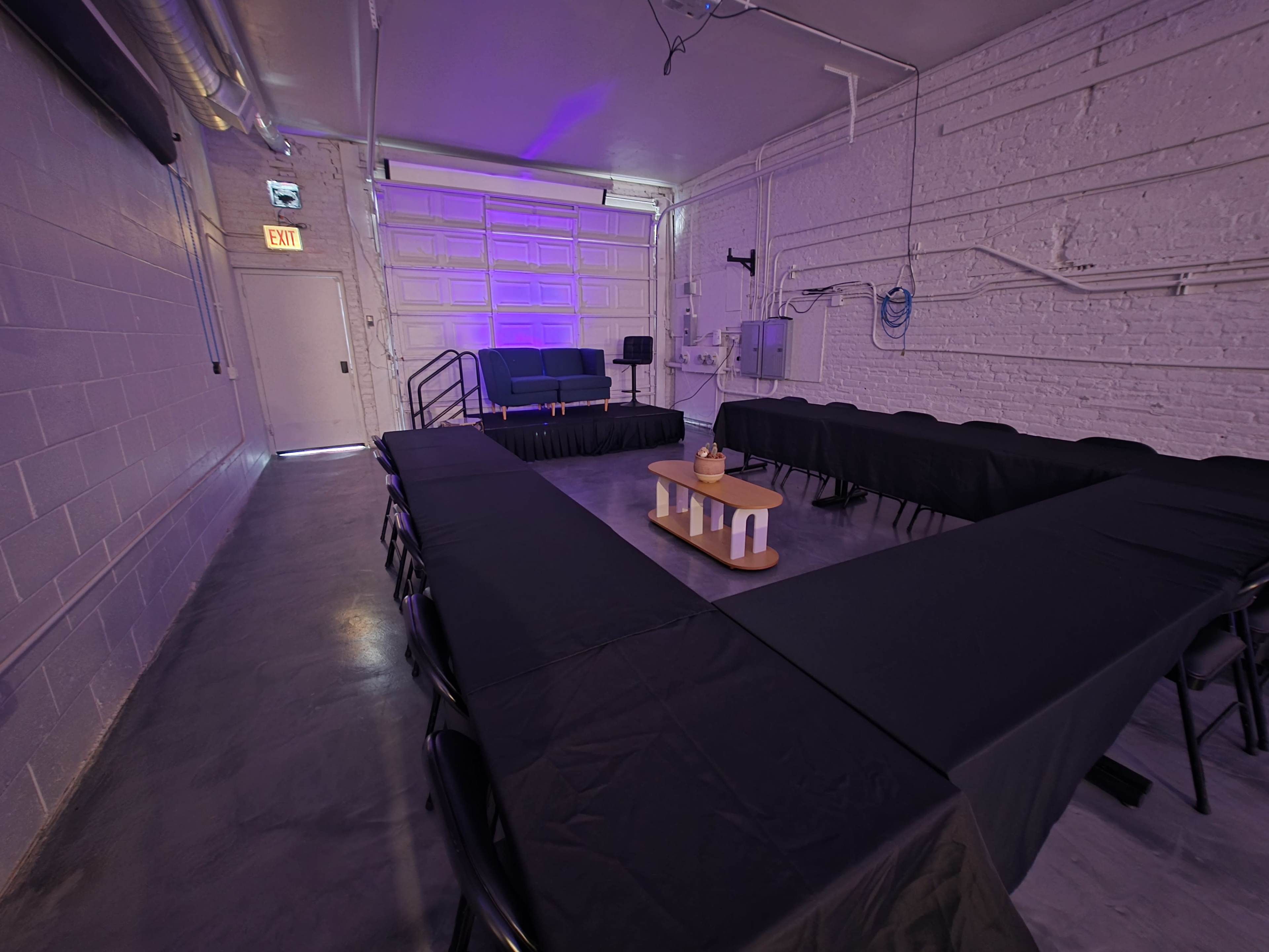 The image shows a spacious room with a rectangular arrangement of black tables and chairs, a small wooden table in the center, and a seating area with a couch against a white brick wall.