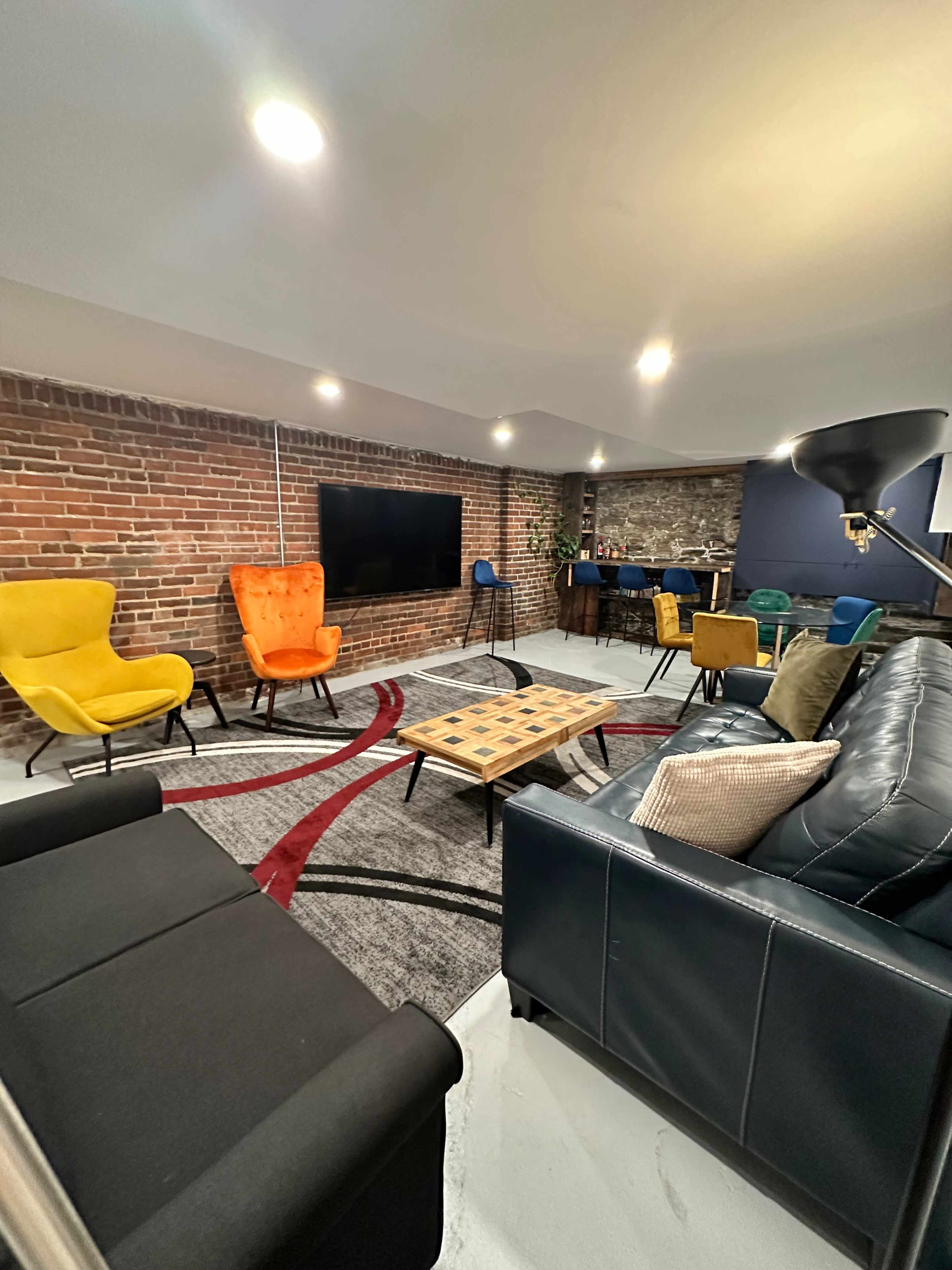 A modern lounge area featuring a mix of colorful chairs, a coffee table, and a large television against a brick wall.
